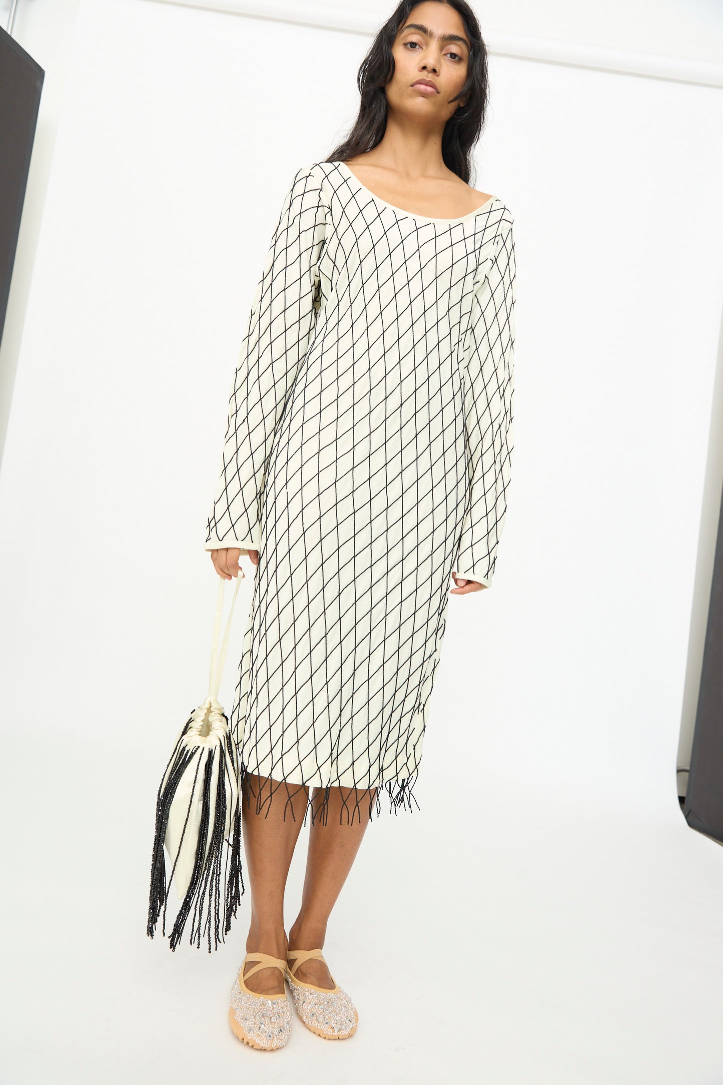 A woman stands against a plain white background, wearing a white dress with a black grid pattern and light-colored shoes, while holding the TIGRA TIGRA Beaded Fringe Wristlet Bag in pale yellow with black beads, handmade in India.