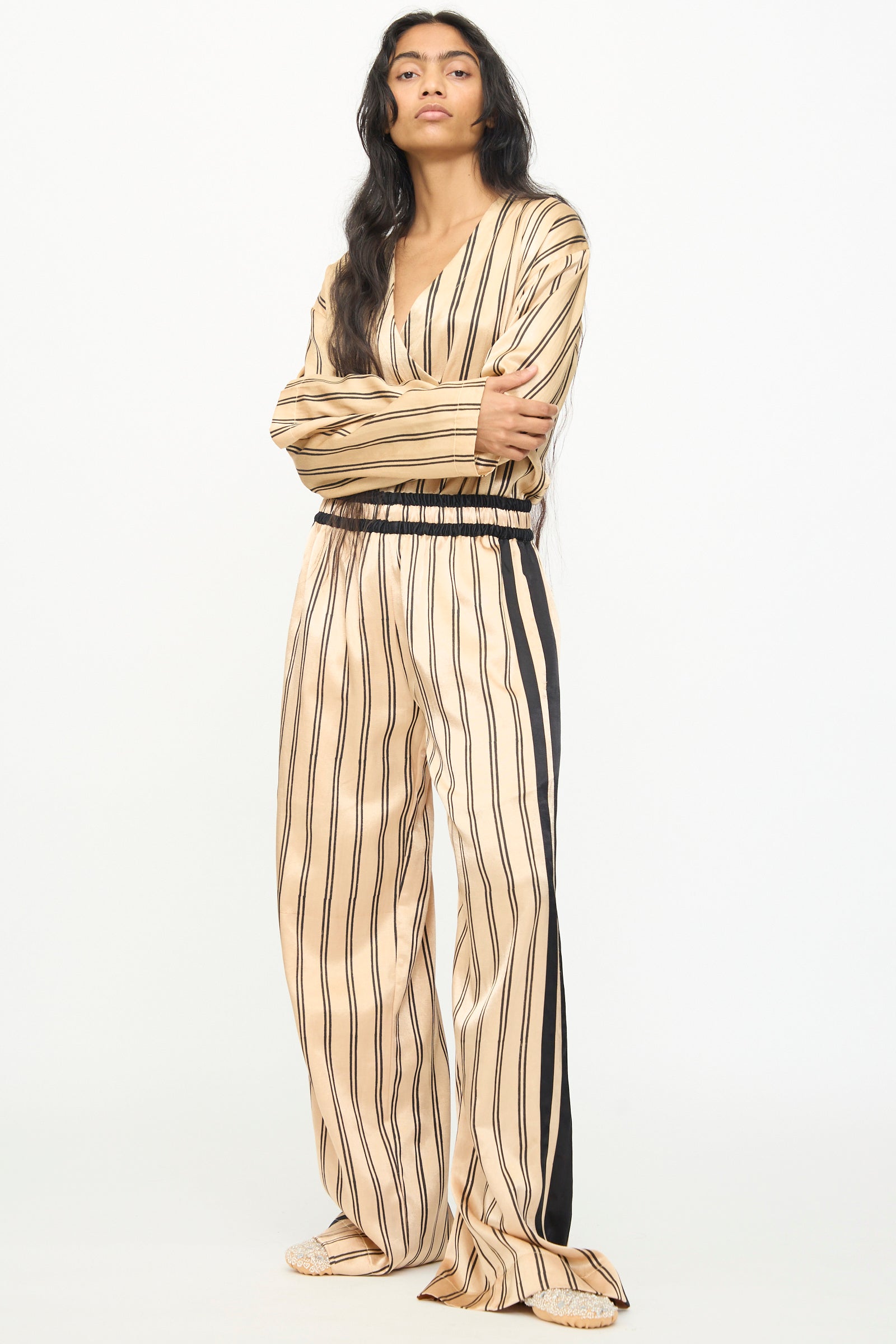 A person stands against a plain background, arms crossed and long, loose hair flowing, wearing TIGRA TIGRA’s Block Printed Boxing Pant in Champagne and Black—crafted from silk mashroo and handmade in India—with a matching beige and black striped long-sleeve top.