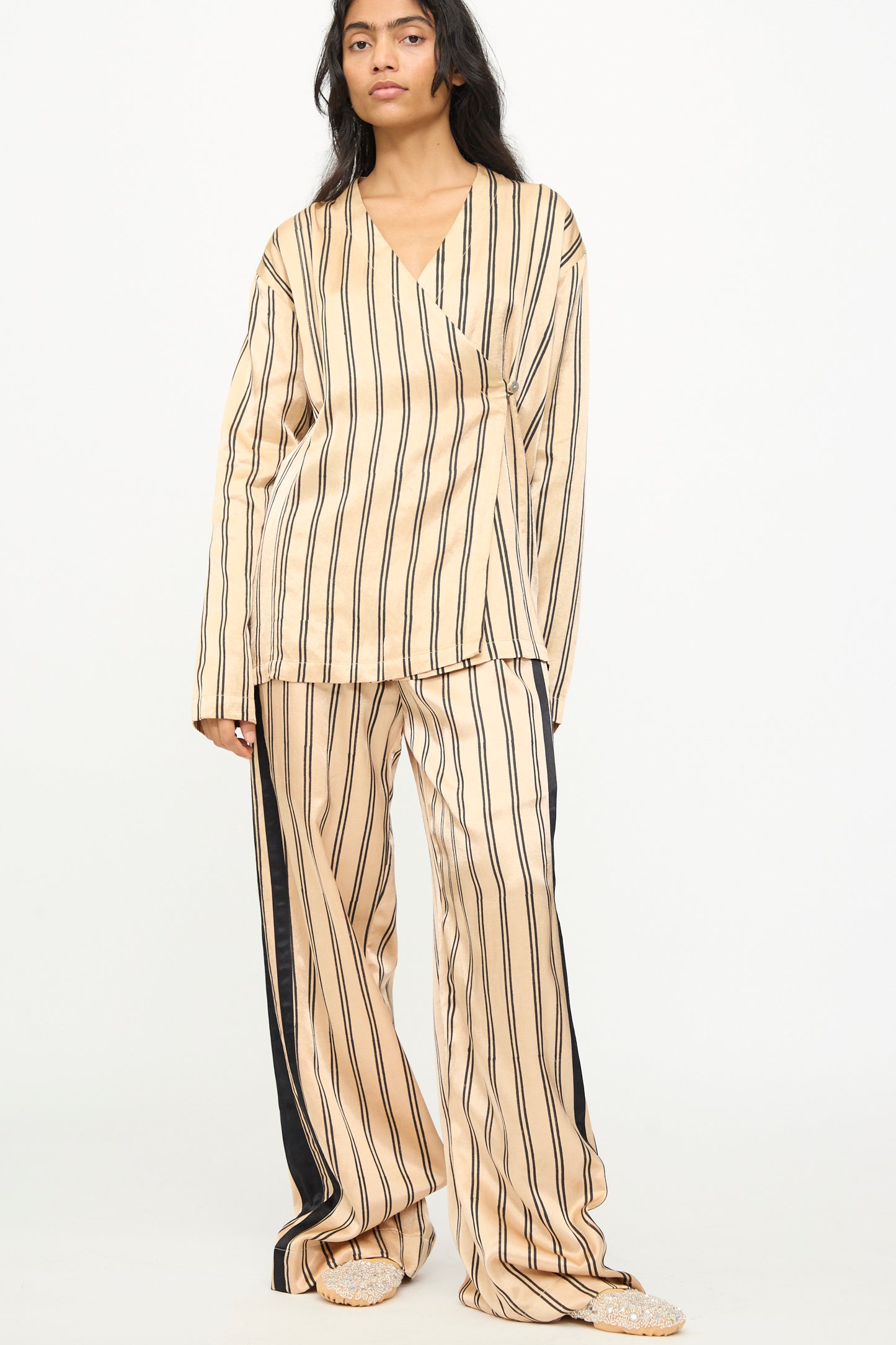 A person stands against a plain white background wearing TIGRA TIGRA's Block Printed Boxing Pant in Champagne and Black, featuring loose-fitting, wide-leg boxing pants with vertical black stripes, long sleeves, and handcrafted from silk mashroo, handmade in India.