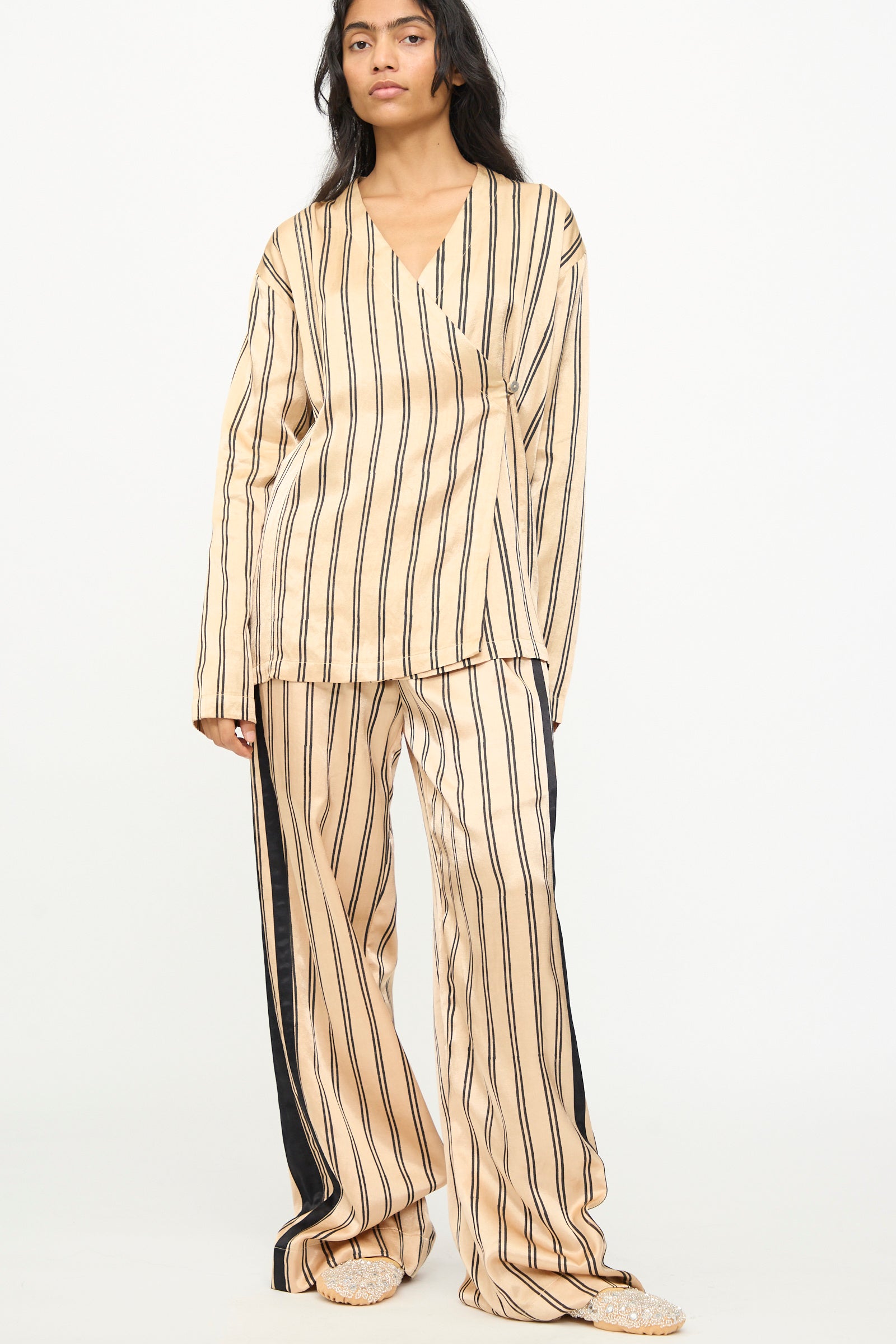 A person stands against a plain white background wearing TIGRA TIGRA's Block Printed Boxing Pant in Champagne and Black, featuring loose-fitting, wide-leg boxing pants with vertical black stripes, long sleeves, and handcrafted from silk mashroo, handmade in India.