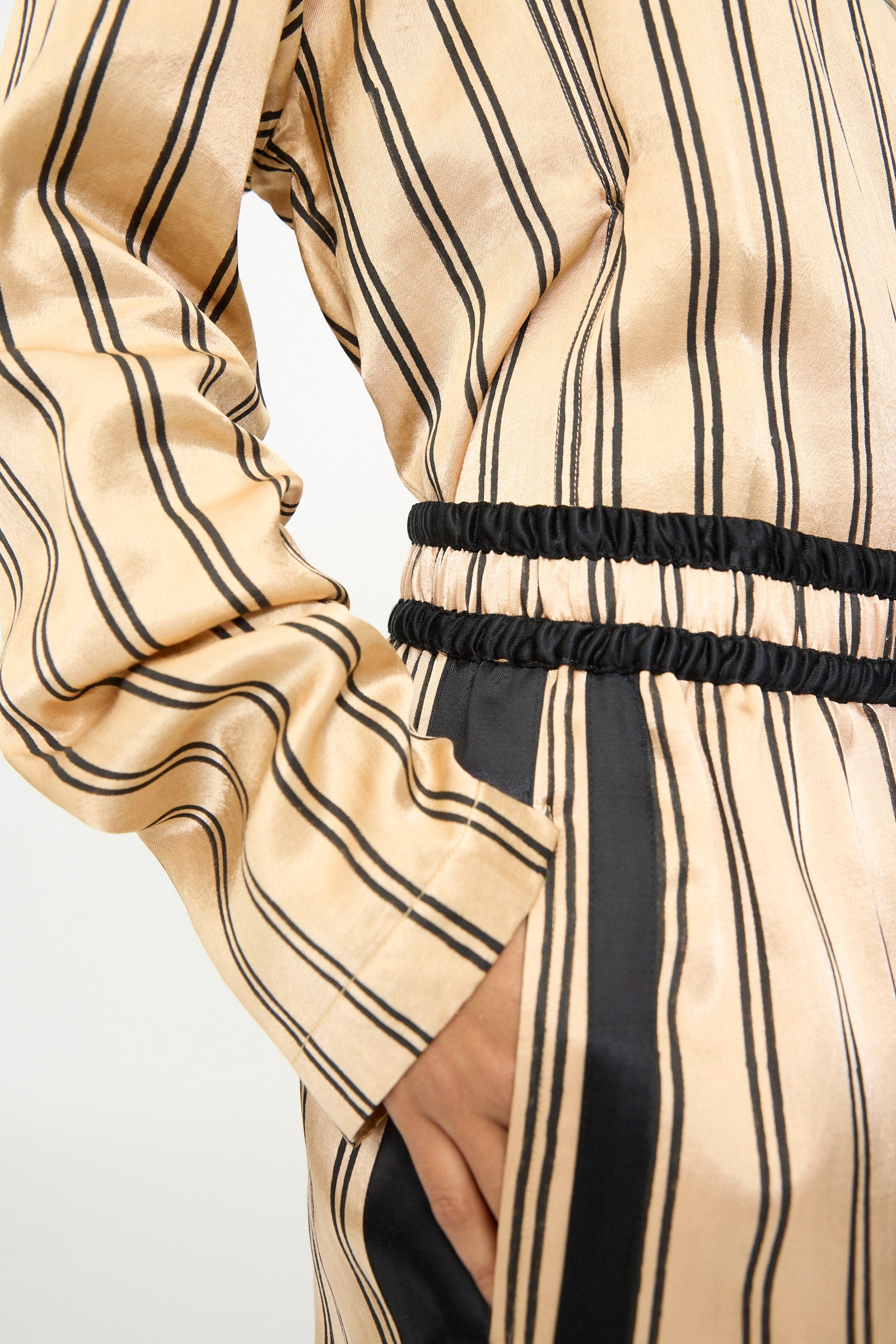 A person wearing the TIGRA TIGRA Block Printed Boxing Pant in Champagne and Black, featuring vertical stripes, an elastic waistband, and pockets, is shown from shoulder to thigh against a plain background. 