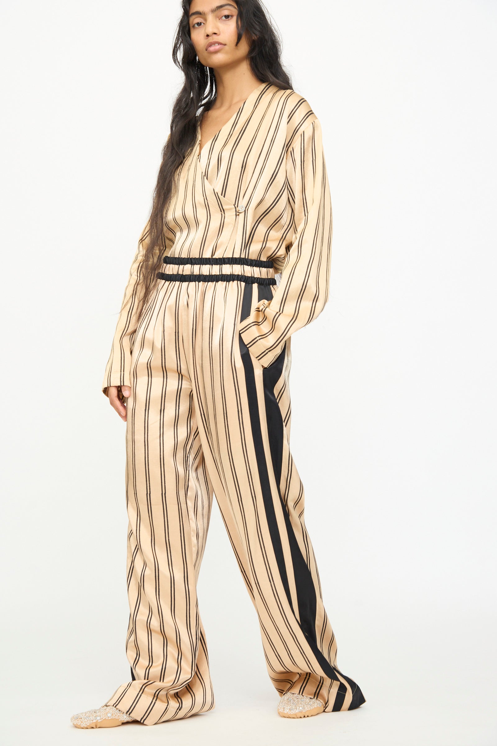 A person stands against a plain white background wearing TIGRA TIGRA's Block Printed Boxing Pant in Champagne and Black—a loose-fitting beige silk mashroo outfit with black vertical stripes, long sleeves, and distinctive black side accents on the handmade pant, crafted in India.