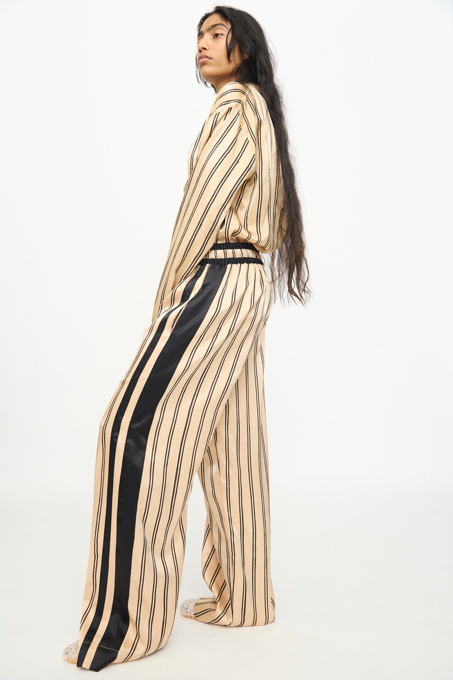 A person stands in profile against a plain white background, wearing TIGRA TIGRA’s Block Printed Boxing Pant in Champagne and Black—a loose-fitting silhouette inspired by classic boxing pants, crafted from silk mashroo with black vertical stripes and a black stripe down the side.