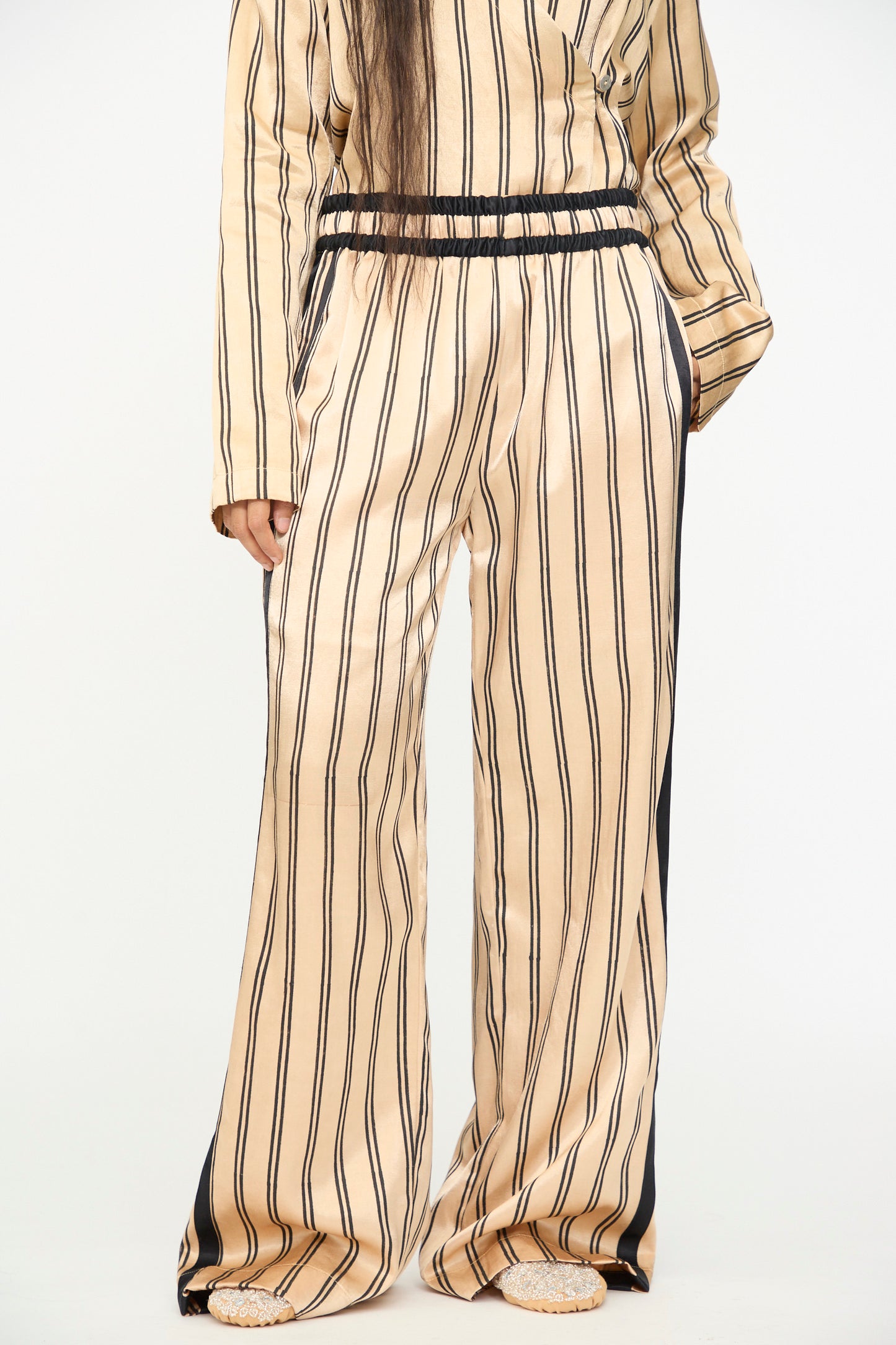 A person wearing TIGRA TIGRA’s Block Printed Boxing Pant in Champagne and Black, paired with a matching vertical striped long-sleeve top, both crafted from luxurious silk mashroo and handmade in India, stands against a plain background.
