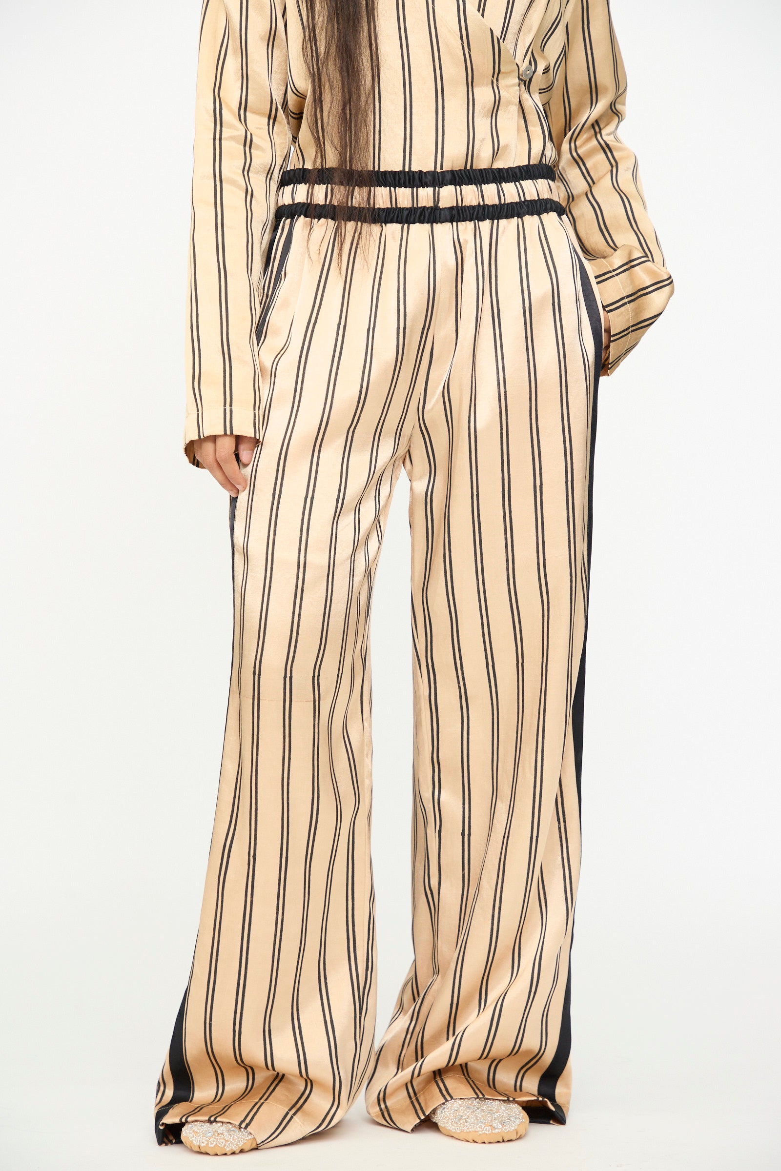 A person wearing TIGRA TIGRA’s Block Printed Boxing Pant in Champagne and Black, paired with a matching vertical striped long-sleeve top, both crafted from luxurious silk mashroo and handmade in India, stands against a plain background.