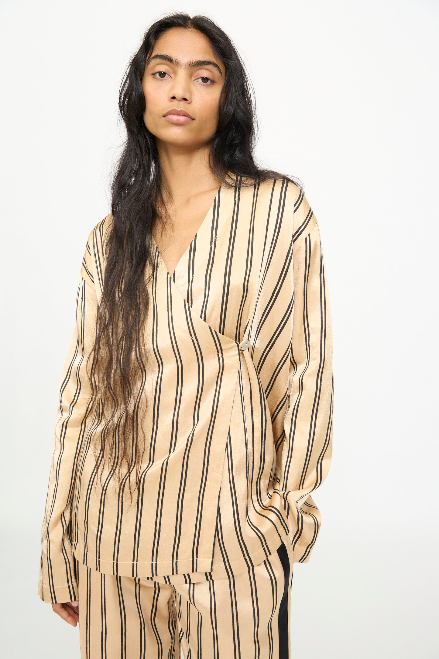 A person with long dark hair is wearing the TIGRA TIGRA Block Printed Striped Wrap Top in Champagne and Black, styled against a plain white background.