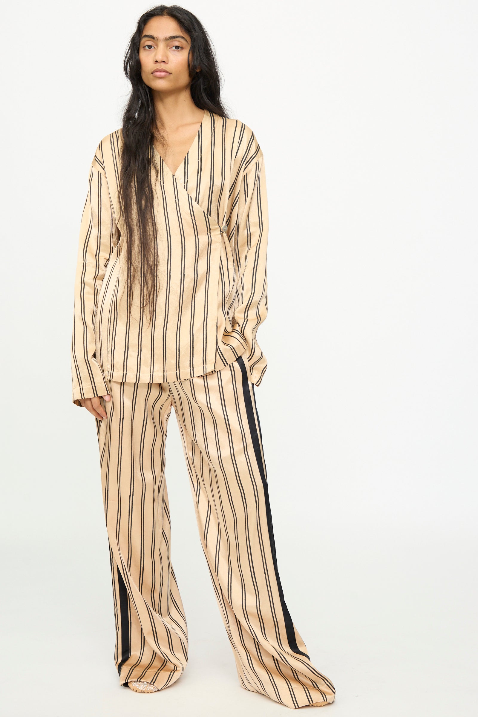 A person stands against a plain background, looking at the camera, dressed in TIGRA TIGRA's Block Printed Striped Wrap Top in Champagne and Black—a beige pajama-style ensemble with vertical black block-printed stripes, long sleeves.