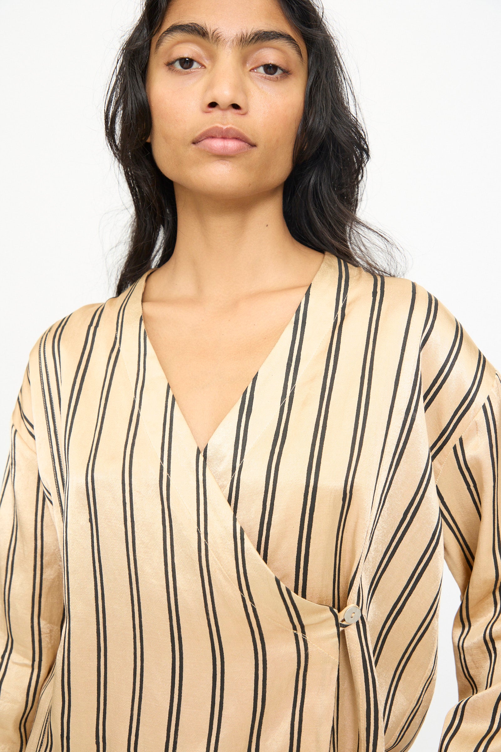 A person with long dark hair wears the TIGRA TIGRA Block Printed Striped Wrap Top in Champagne and Black, a hand-loomed silk kimono-style blouse featuring black block printed stripes and a v-neck, standing against a plain background.