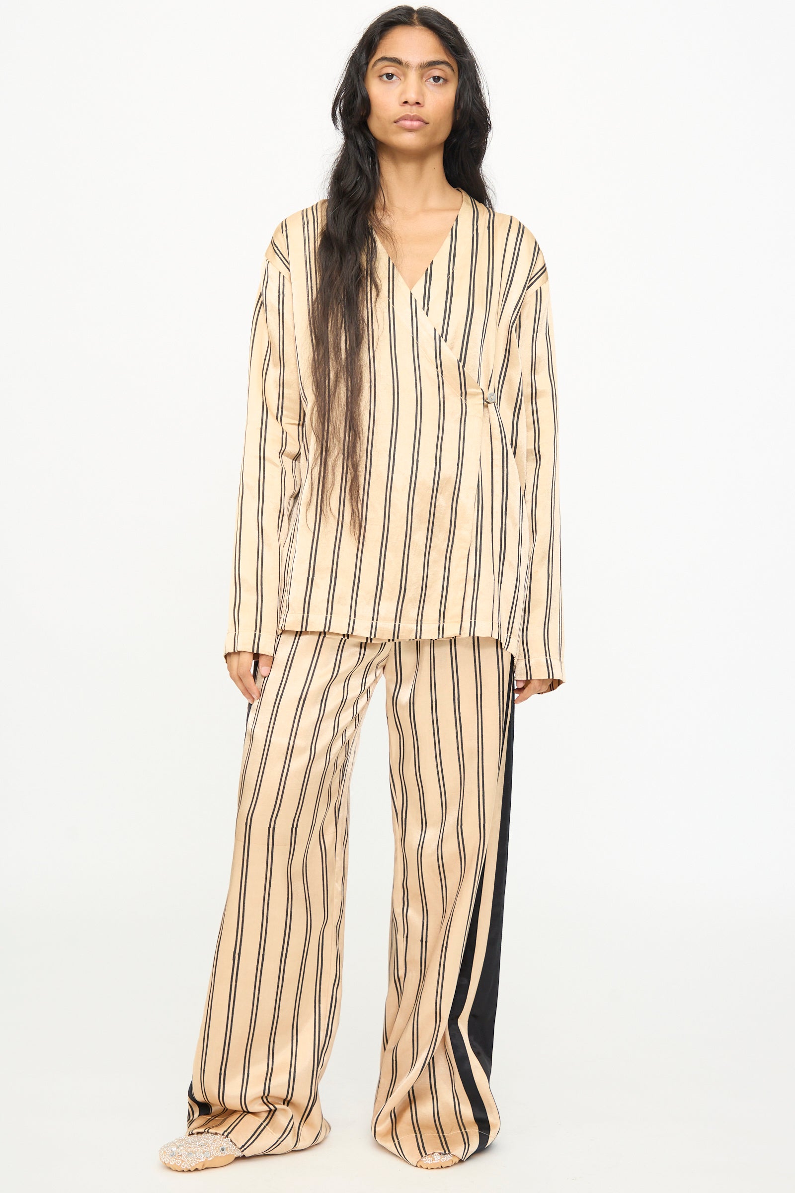 A person stands facing forward wearing TIGRA TIGRA's Block Printed Striped Wrap Top in Champagne and Black, crafted from hand-loomed silk with block printed stripes, set against a plain white background.