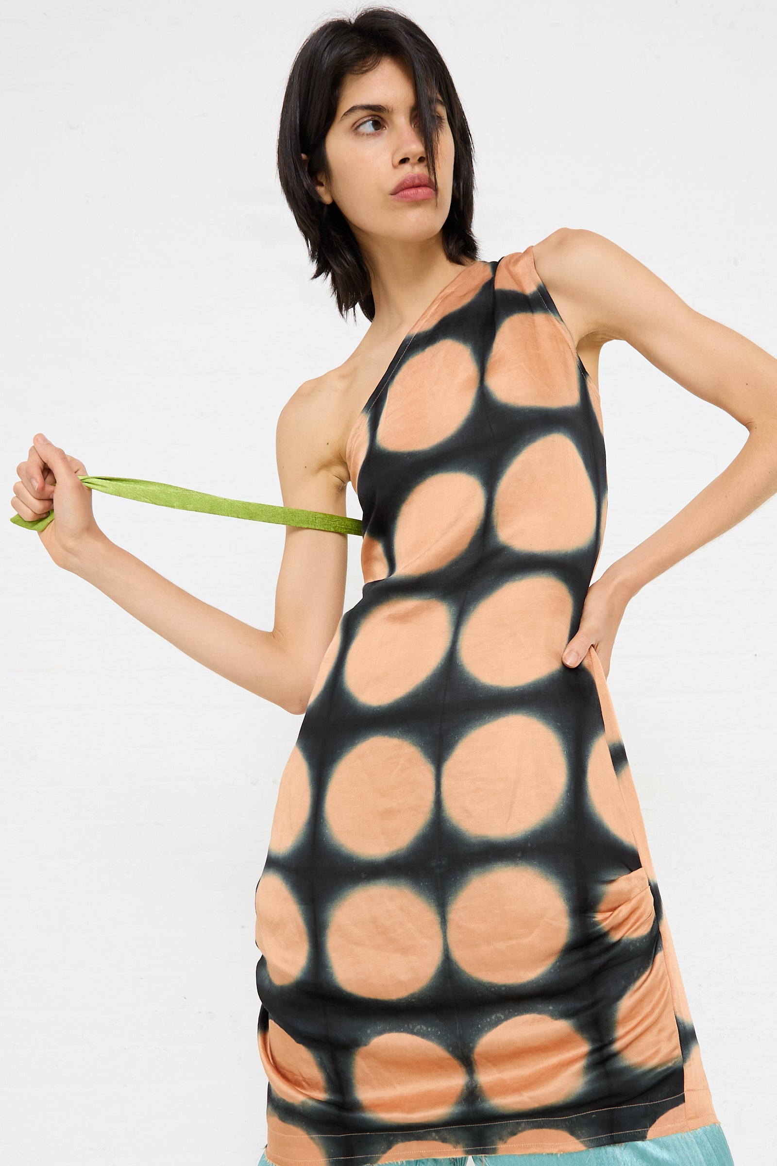 A person wearing a TIGRA TIGRA One Shoulder Mini Dot Dress holds a green strap while looking to the side. The background is plain white.