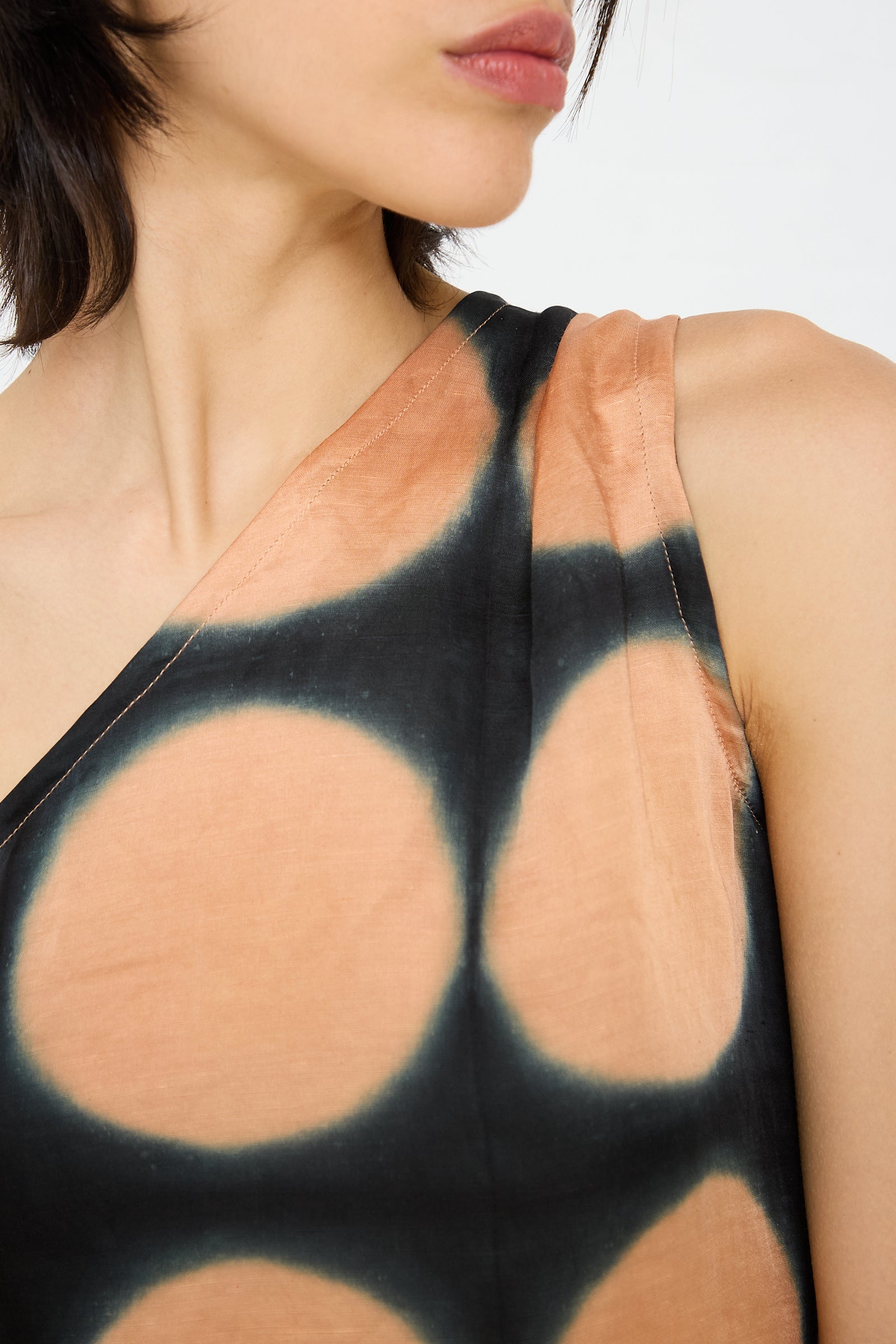 Close-up of a person wearing the TIGRA TIGRA One Shoulder Mini Dot Dress. The fabric features a hand-dyed tie-dye design in black and peach. The person's face is partially visible, looking away from the camera.