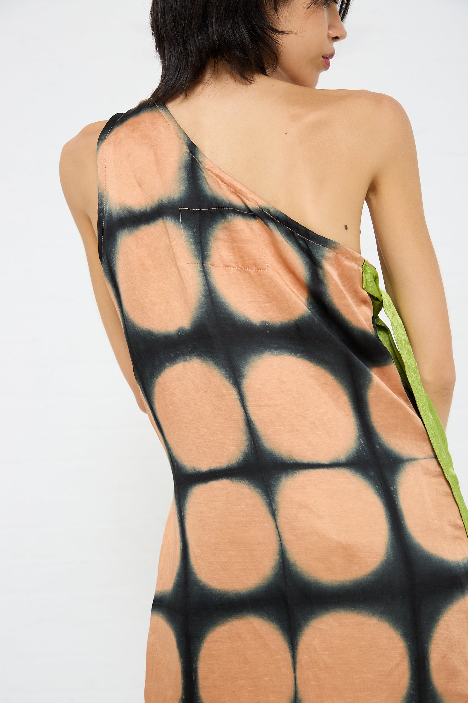A person wearing the TIGRA TIGRA One Shoulder Mini Dot Dress, a sleeveless, asymmetrical, hand-dyed dress with a circular pattern design in black and peach. The linen/silk blend dress features a green stripe along the side.