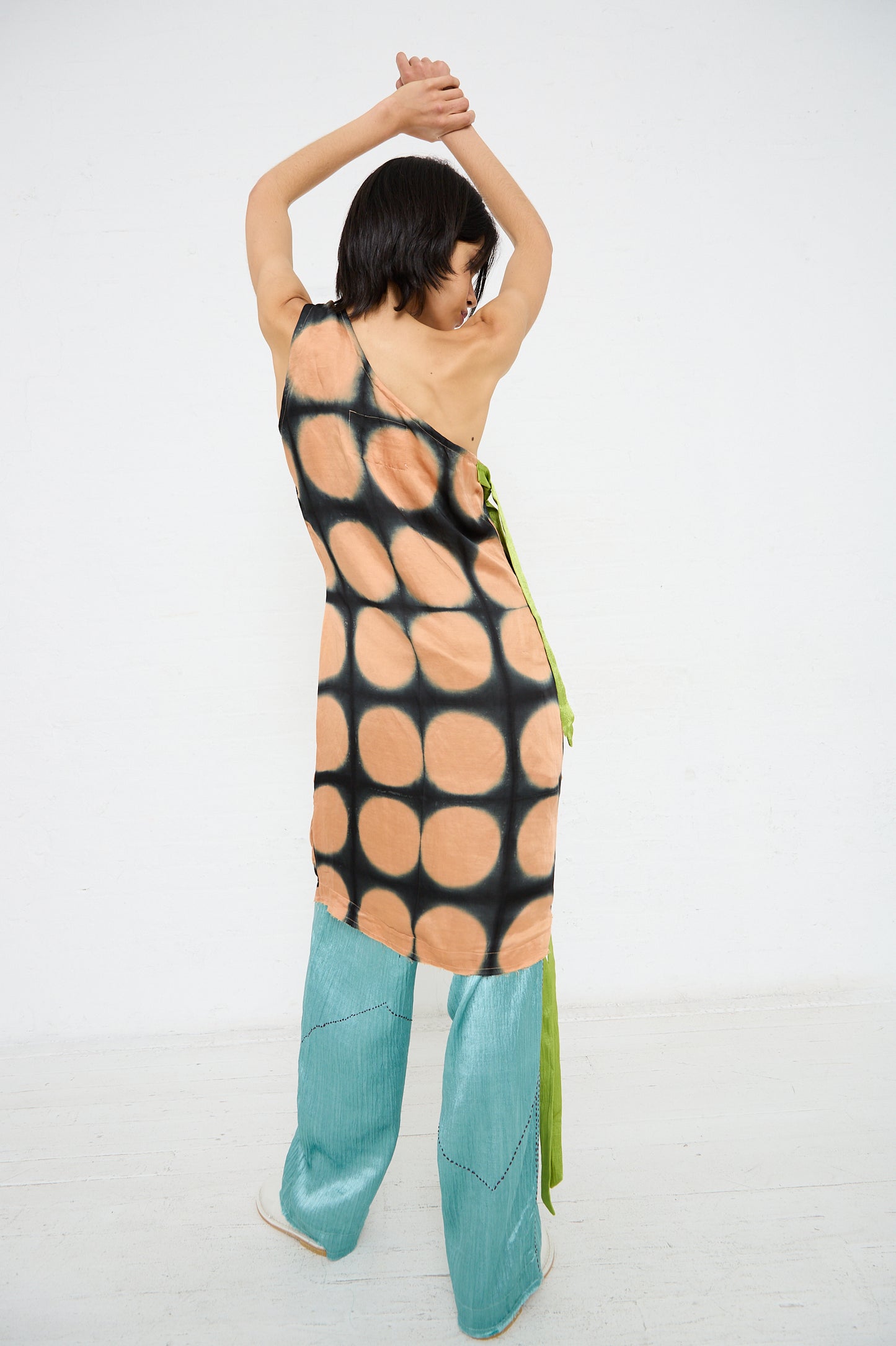 A person stands with their back to the camera, arms raised, wearing a sleeveless One Shoulder Mini Dot Dress by TIGRA TIGRA and blue pants. The background is a plain white wall and floor.