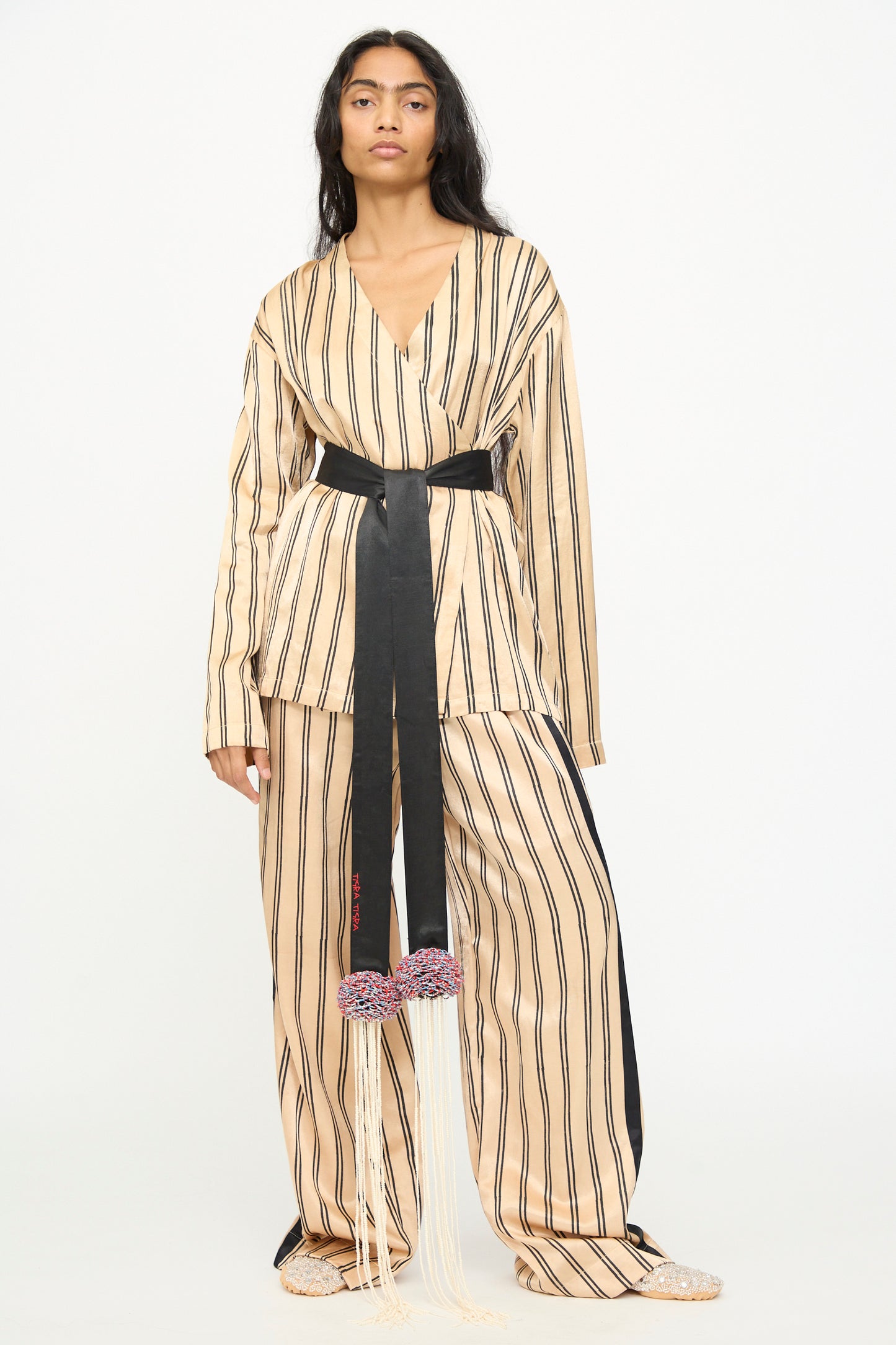 A person stands against a plain white background wearing wide pants and a matching top in beige and black vertical stripes, styled with the TIGRA TIGRA Pillows Belt with Glass Bead Fringe—a hand-loomed silk belt adorned with decorative tassels and beaded fringe—and paired with beige shoes.