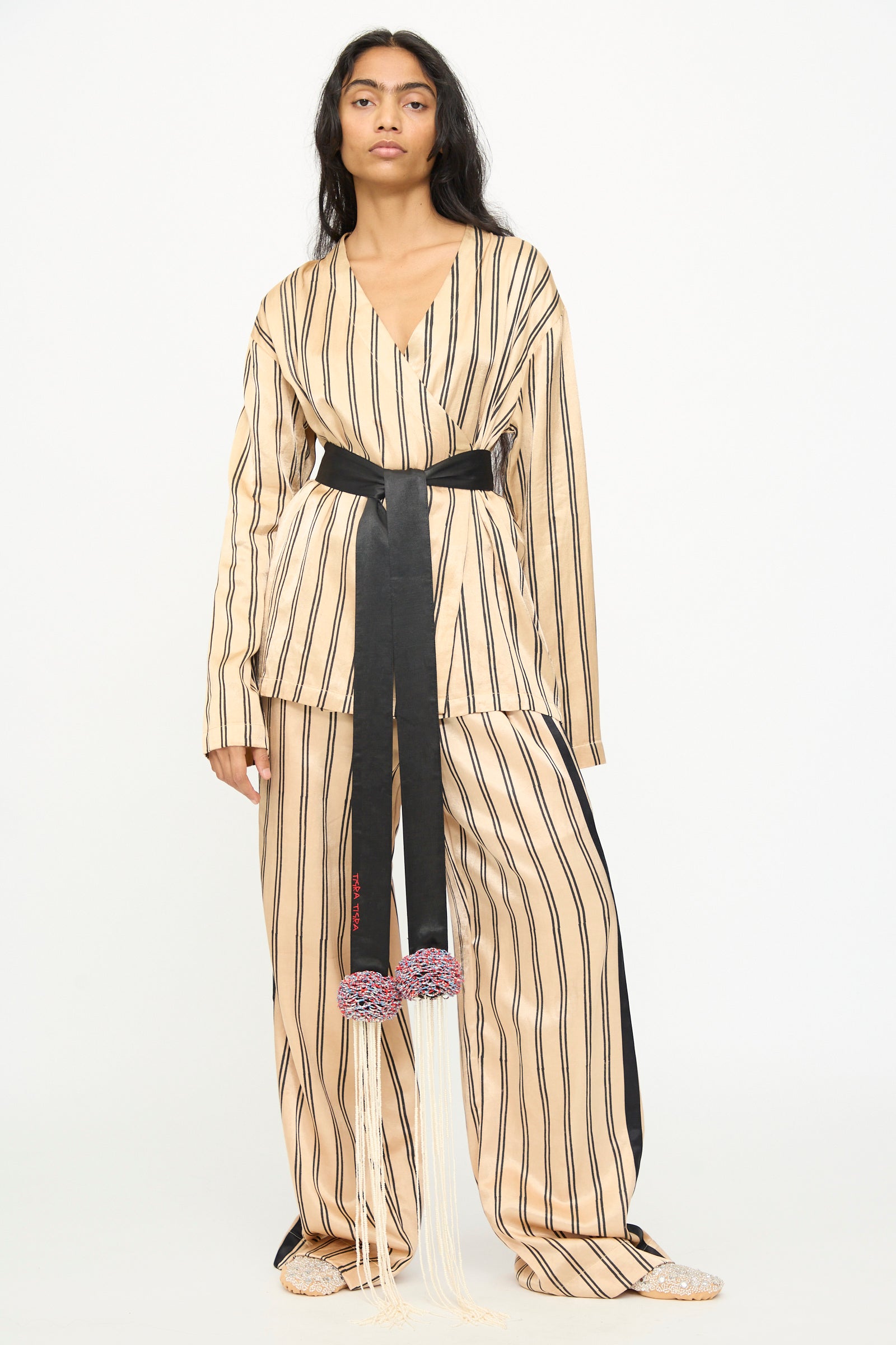 A person stands against a plain white background wearing wide pants and a matching top in beige and black vertical stripes, styled with the TIGRA TIGRA Pillows Belt with Glass Bead Fringe—a hand-loomed silk belt adorned with decorative tassels and beaded fringe—and paired with beige shoes.