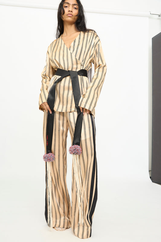A person wearing a beige outfit with vertical black stripes and wide-leg pants is styled with the TIGRA TIGRA Pillows Belt with Glass Bead Fringe, handmade in India, standing against a plain white background.
