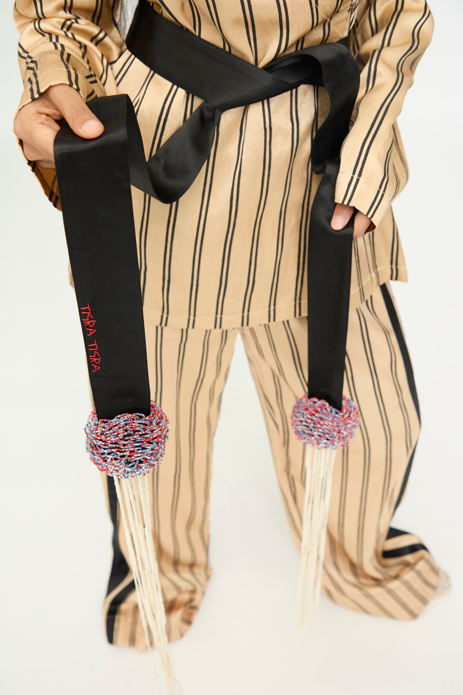 A person in a tan and black striped outfit holds the TIGRA TIGRA Pillows Belt with Glass Bead Fringe, a hand-loomed silk belt handmade in India, featuring colorful braided tassels, white fringe, and glass bead accents at the ends, standing against a white background.