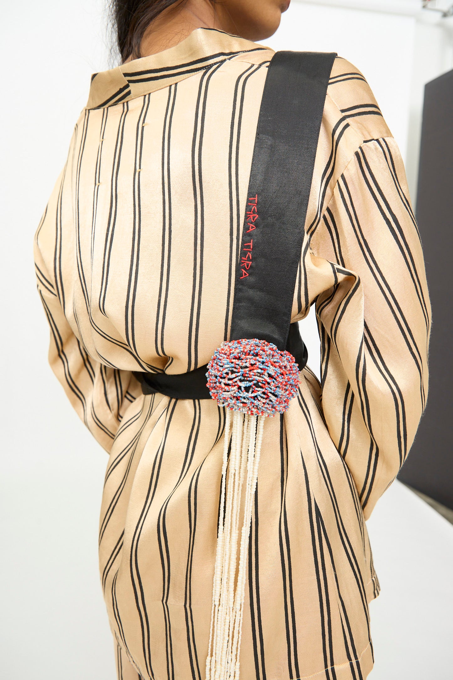 Person wearing a beige and black striped outfit with a black sash embroidered with "TIGRA TIGRA"—the Pillows Belt with Glass Bead Fringe, a stunning, handmade in India beaded belt featuring a circular ornament and fringe hanging at the back.