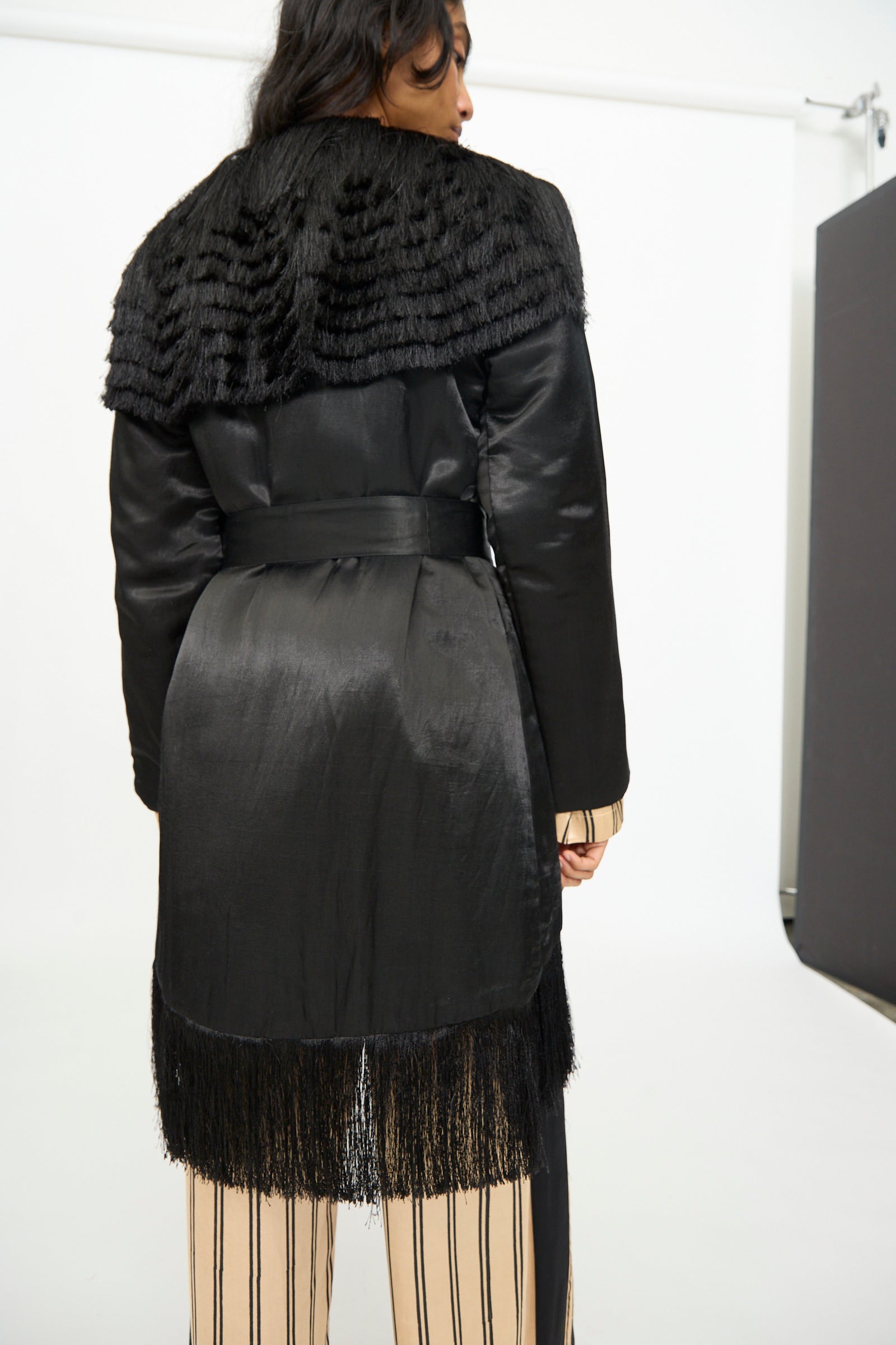 A person photographed from the back wears the TIGRA TIGRA Puffy Silk Fringe Wrap Coat in Black, featuring a textured fur-like collar and fringe hem, layered over a beige and black striped outfit.