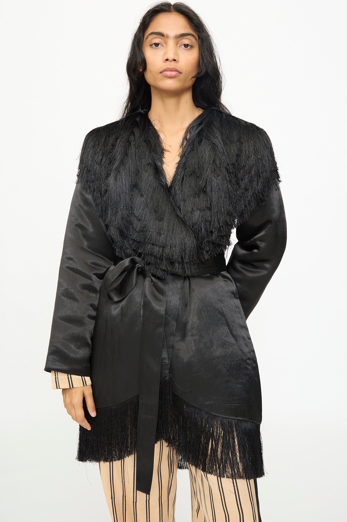 Wearing the TIGRA TIGRA Puffy Silk Fringe Wrap Coat in Black—a handmade, belted jacket with fringe details from India—over a beige outfit with black vertical stripes, the person stands against a plain white background.