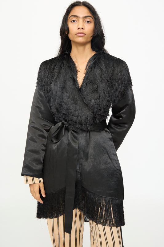 Wearing the TIGRA TIGRA Puffy Silk Fringe Wrap Coat in Black—a handmade, belted jacket with fringe details from India—over a beige outfit with black vertical stripes, the person stands against a plain white background.