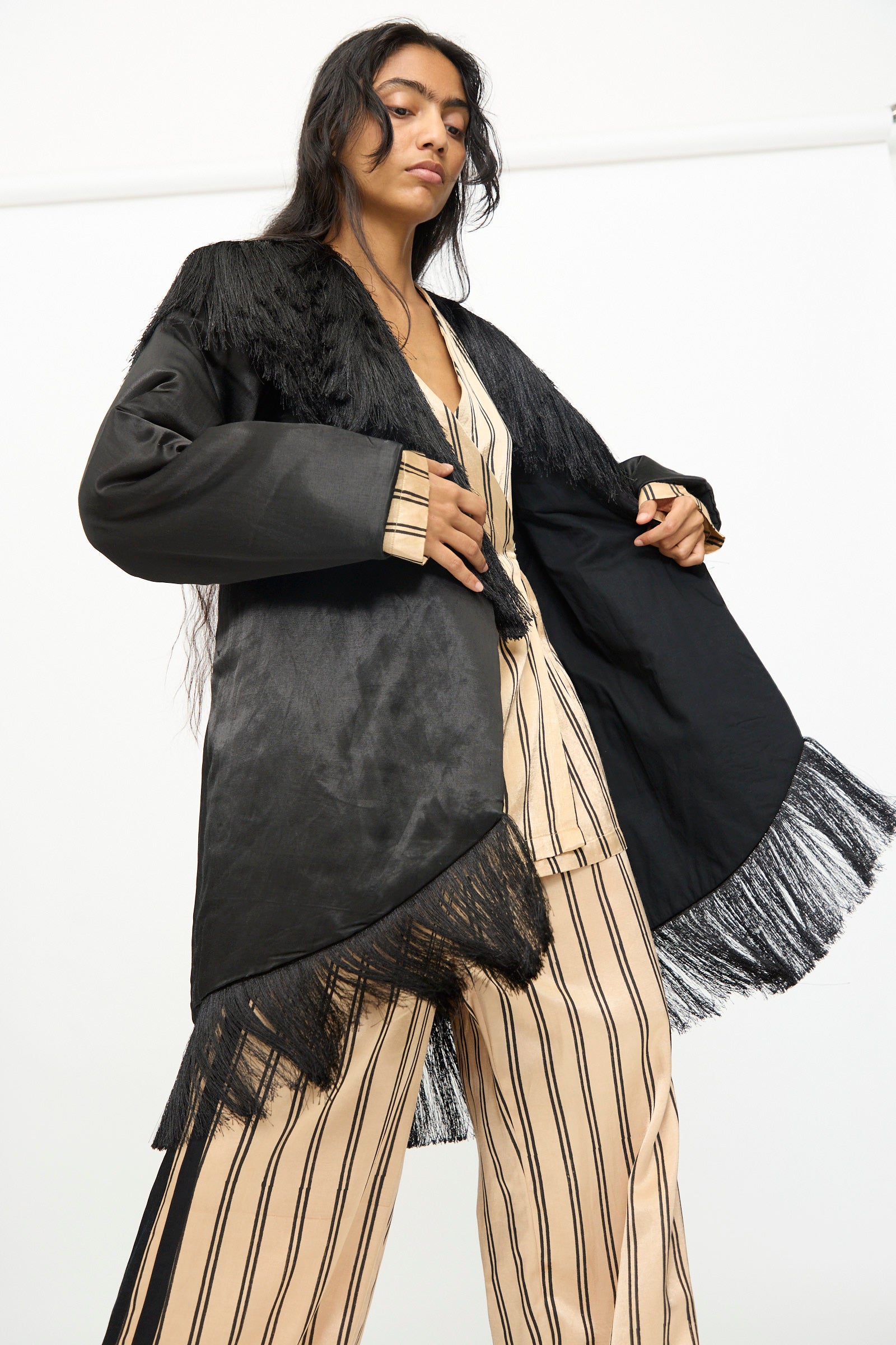 A woman wearing the TIGRA TIGRA Puffy Silk Fringe Wrap Coat in Black over striped beige pajamas, standing against a plain white background.