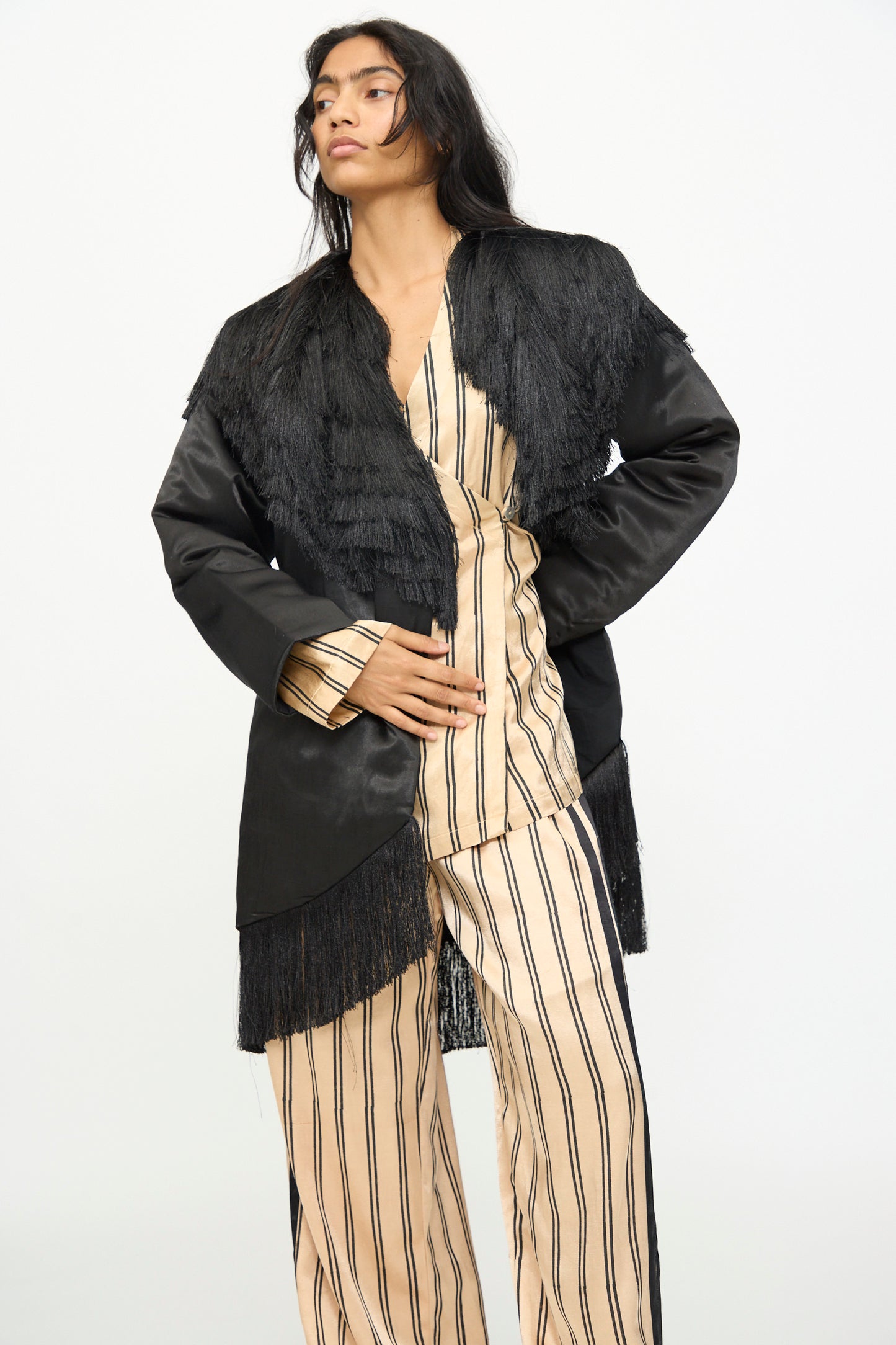 Draped in the TIGRA TIGRA Puffy Silk Fringe Wrap Coat in Black, a person stands against a plain white background, layering the handmade-in-India statement piece over a beige outfit with black vertical stripes.