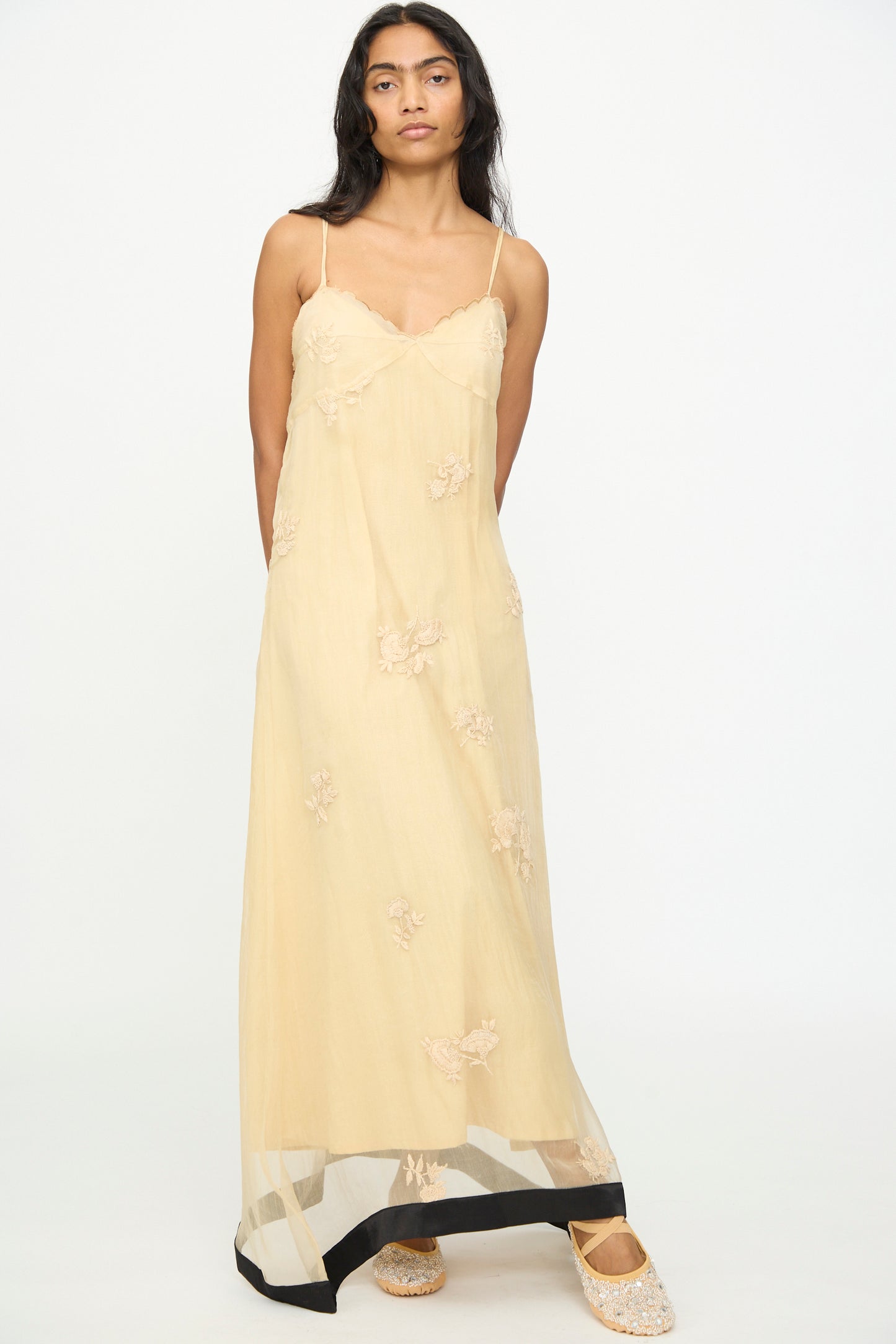 A woman stands against a plain background wearing TIGRA TIGRA’s Sheer Nightime Dress with Hand Embroidery in Sand with Black— a long, sheer, light beige silk chanderi slip dress featuring thin straps, hand-embroidered flowers, and black trim at the hem—paired with embellished flat shoes.