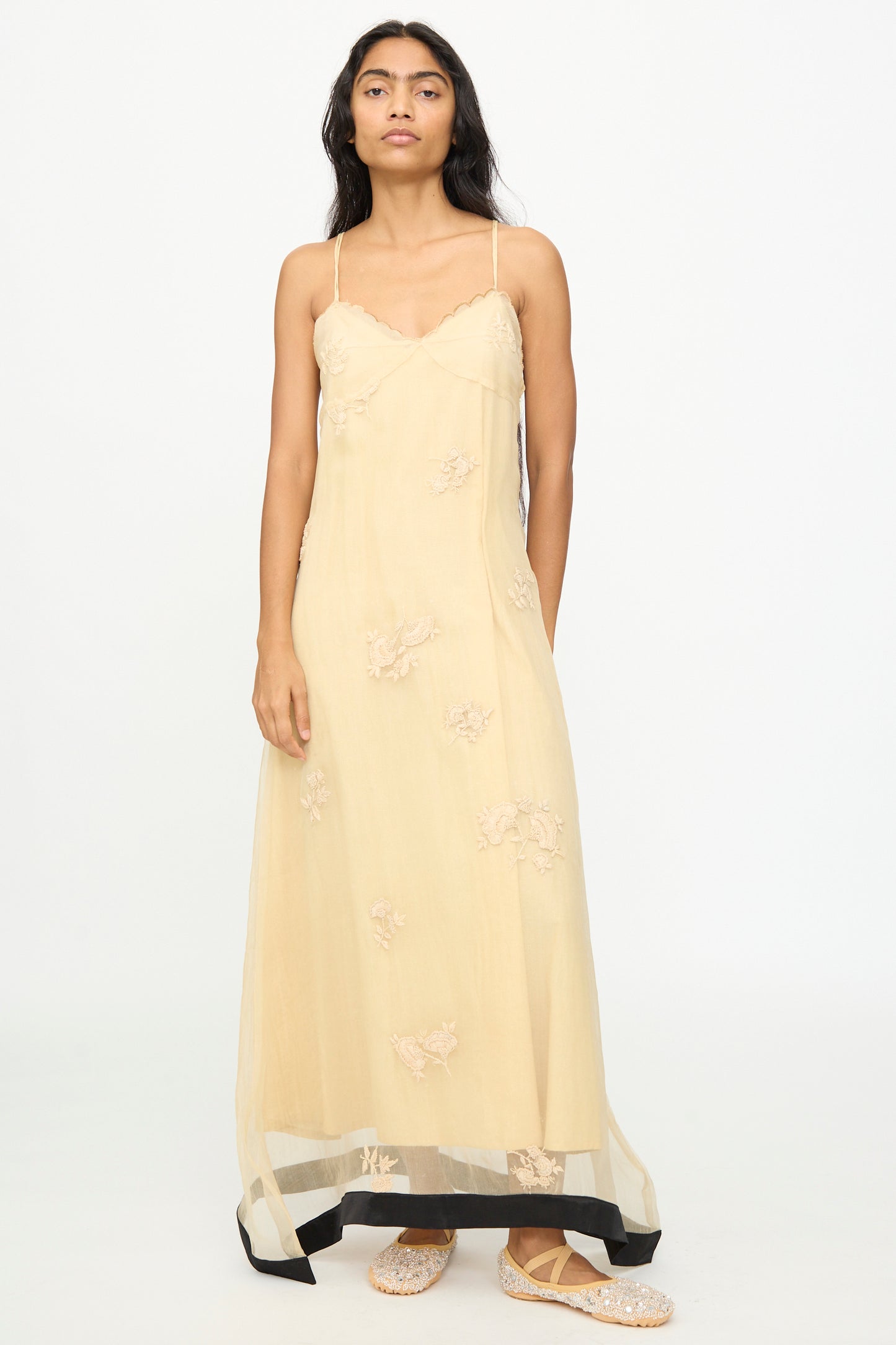 A woman stands against a plain white background, wearing the TIGRA TIGRA Sheer Nightime Dress with Hand Embroidery in Sand with Black—a long, pale yellow silk Chanderi slip dress featuring thin straps and delicate hand-embroidered flowers—paired with flat, textured shoes. 