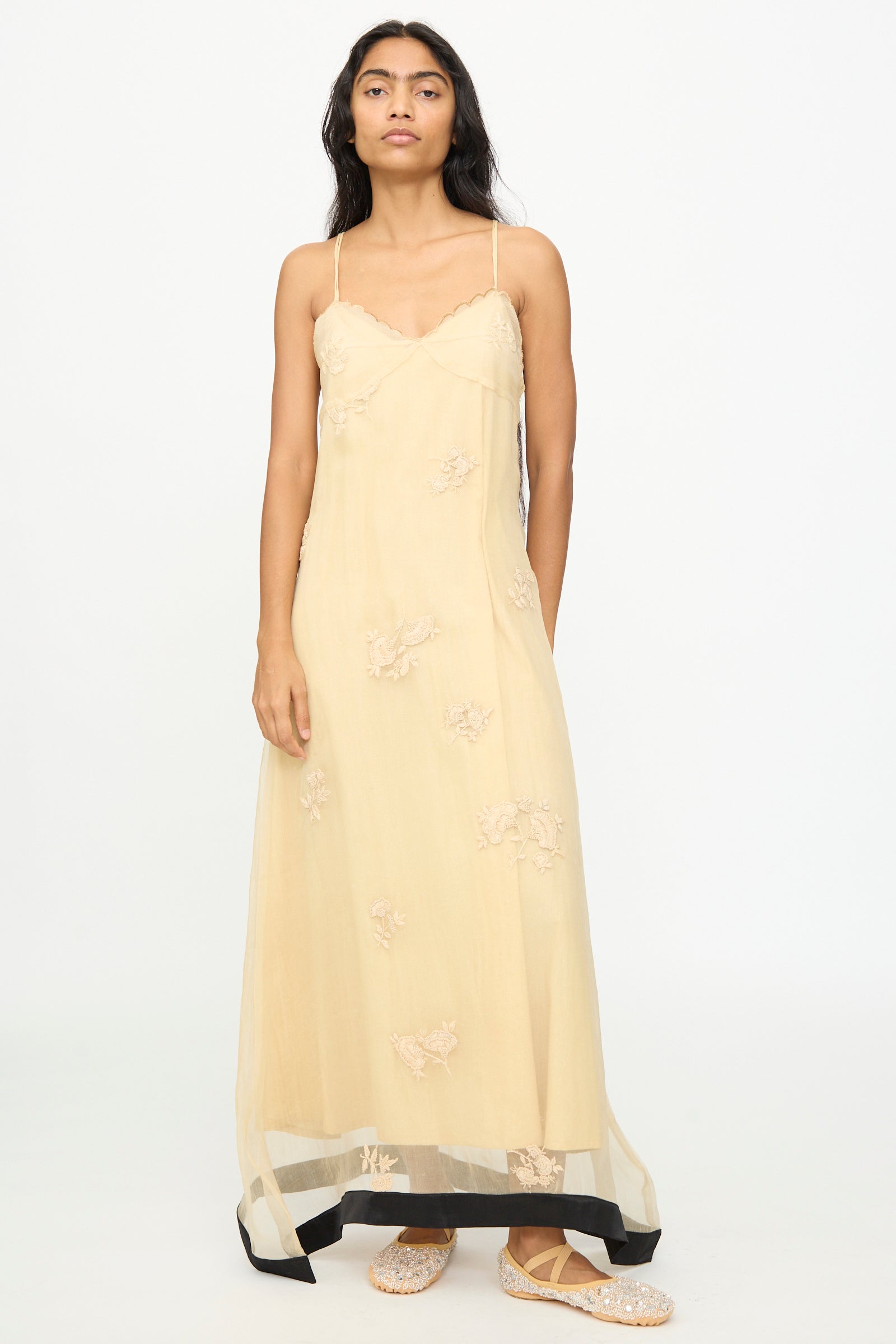A woman stands against a plain white background, wearing the TIGRA TIGRA Sheer Nightime Dress with Hand Embroidery in Sand with Black—a long, pale yellow silk Chanderi slip dress featuring thin straps and delicate hand-embroidered flowers—paired with flat, textured shoes. 