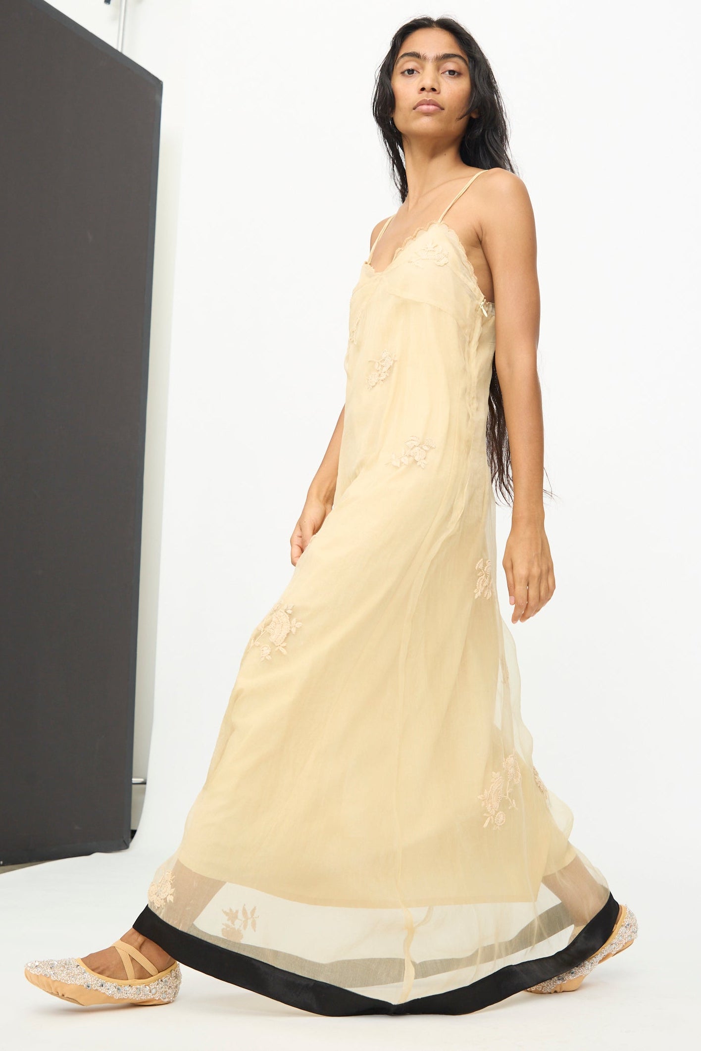 A woman walks in a studio with a white background, wearing the TIGRA TIGRA Sheer Nightime Dress with Hand Embroidery in Sand with Black.