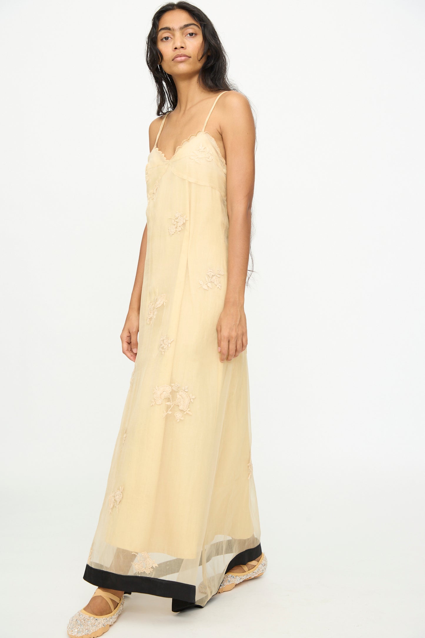 A woman stands against a white background, wearing the TIGRA TIGRA Sheer Nightime Dress with Hand Embroidery in Sand with Black, featuring a long, sheer silhouette, thin straps, and hand-embroidered flowers, paired with embroidered flats.