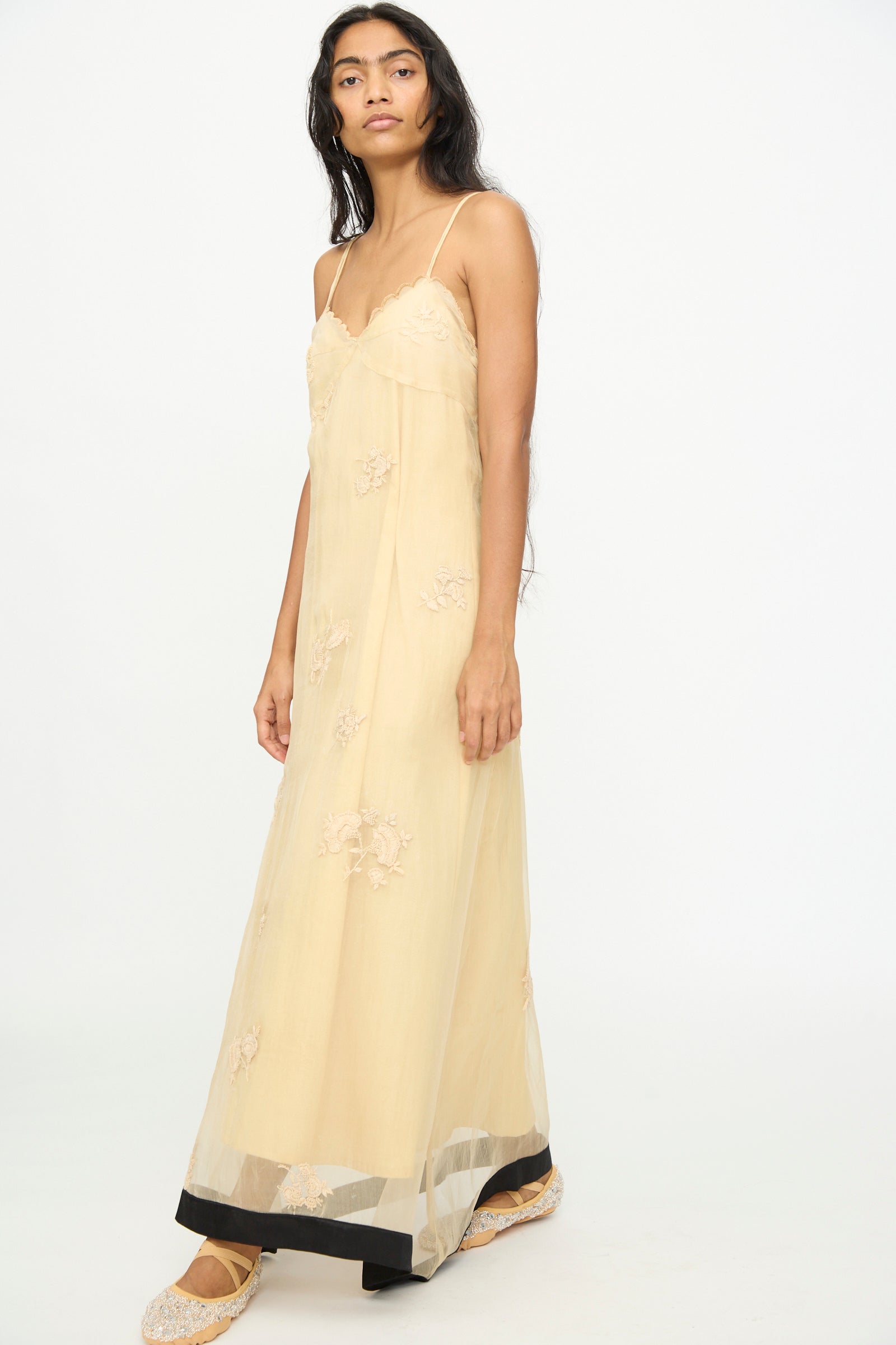 A woman stands against a white background, wearing the TIGRA TIGRA Sheer Nightime Dress with Hand Embroidery in Sand with Black, featuring a long, sheer silhouette, thin straps, and hand-embroidered flowers, paired with embroidered flats.