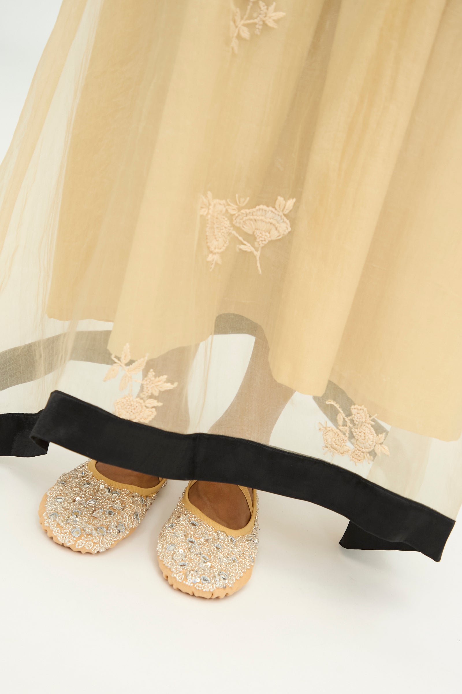 Close-up of TIGRA TIGRA’s Sheer Nightime Dress with Hand Embroidery in Sand with Black—a beige silk chanderi slip dress featuring hand-embroidered flowers and a black border—revealing a pair of feet in tan, beaded shoes against a white background. 