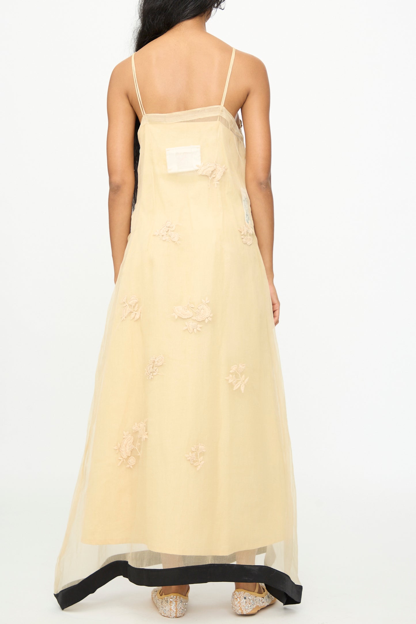 A woman stands facing away against a plain white background, wearing the TIGRA TIGRA Sheer Nightime Dress with Hand Embroidery in Sand with Black—a sleeveless beige silk chanderi slip dress featuring hand-embroidered floral details and a contrasting black hem.