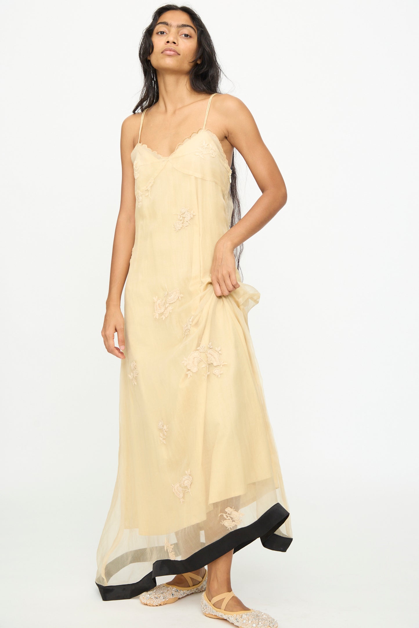 A woman stands on a plain background, holding one side of her Sheer Nightime Dress with Hand Embroidery in Sand with Black by TIGRA TIGRA. 