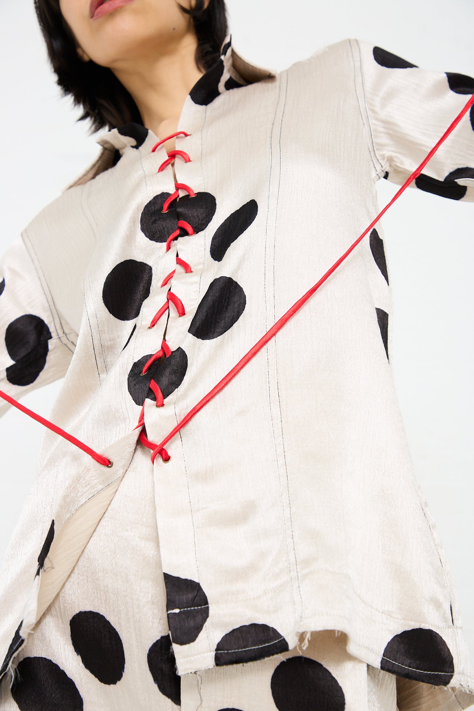 Person wearing a light-colored, long sleeve Silk Mashroo Lace Up Shirt in White by TIGRA TIGRA with large black polka dots and a red lace-up detail down the front. The person’s face is partially visible, and the background is plain white.