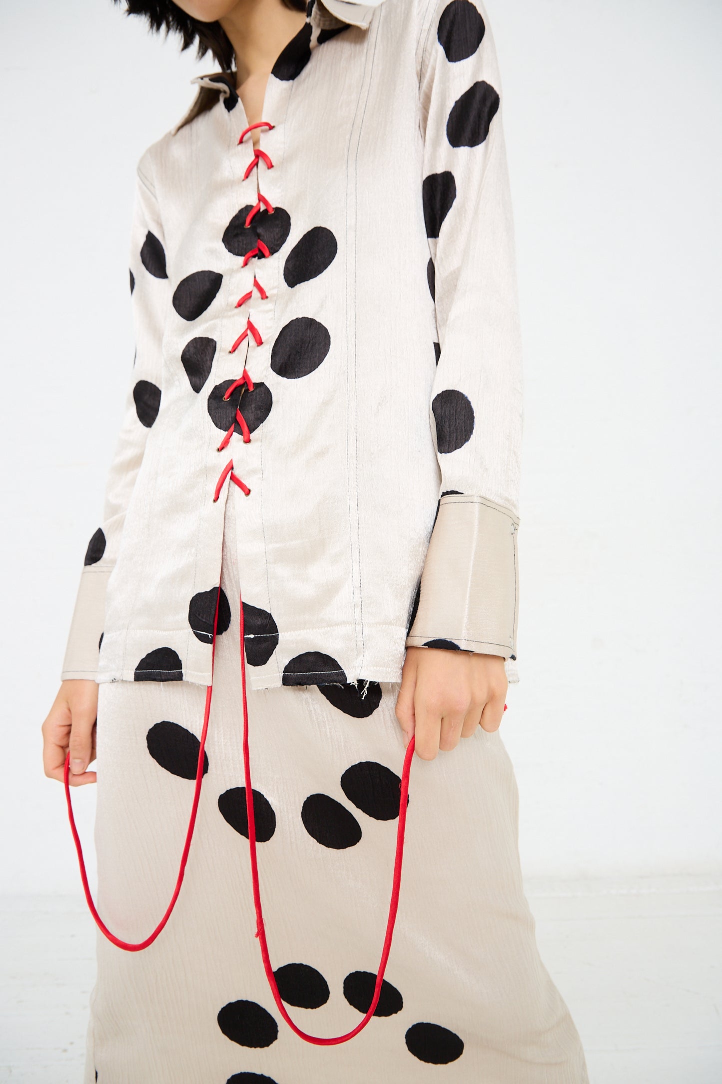 Person wearing a cream-colored, black polka-dotted outfit, featuring the Silk Mashroo Lace Up Shirt in White by TIGRA TIGRA with red lace-up detail and matching skirt, holding red cords hanging from the front. The unique pattern appears almost as hand clamp dyed moon dots. Face is partially out of frame.