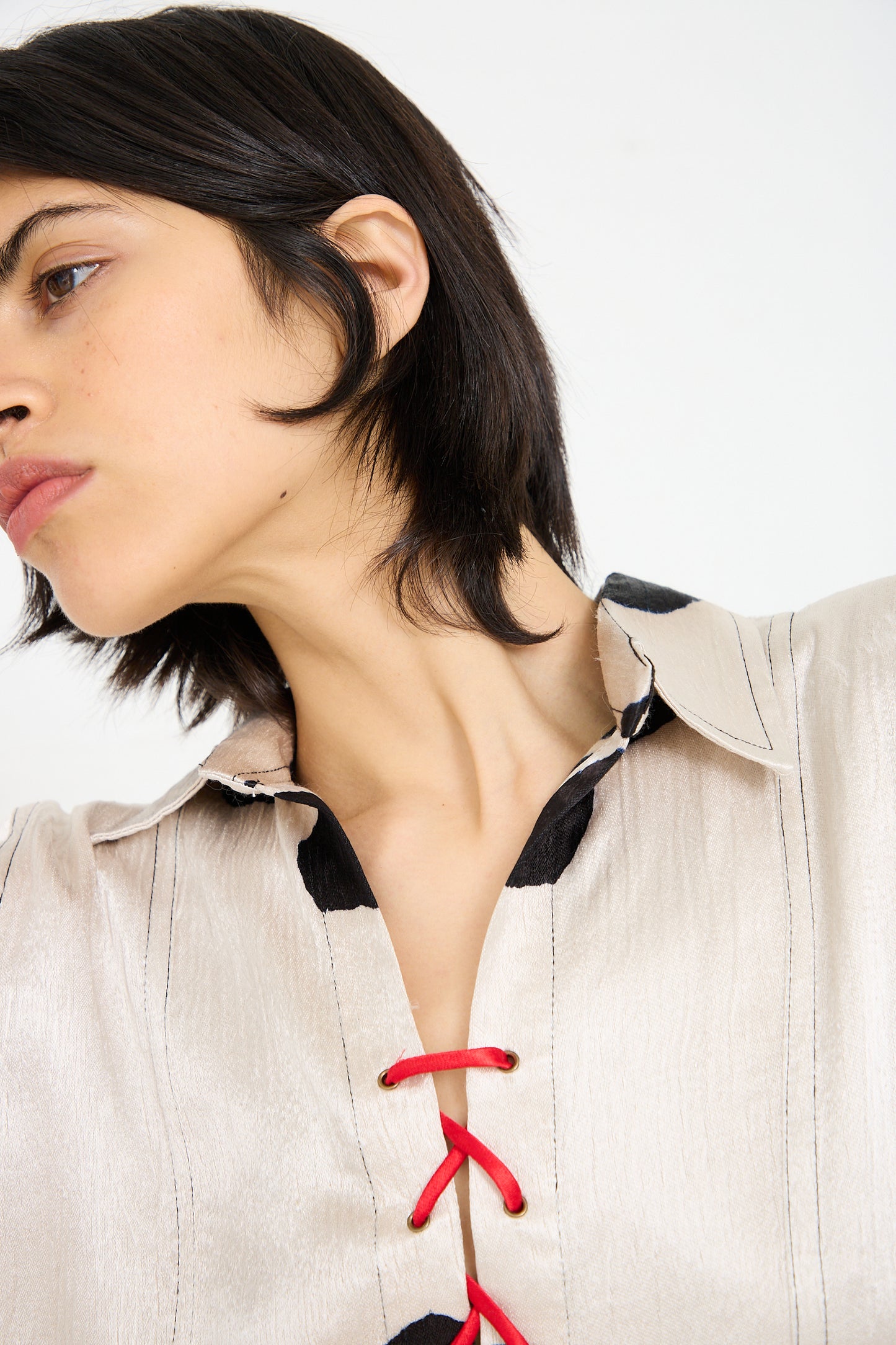 A person with short dark hair is shown in a close-up wearing a Silk Mashroo Lace Up Shirt in White by TIGRA TIGRA, which is a light-colored, long sleeve lace-up shirt with red detailing and black accents. The person's head is slightly turned to the side.