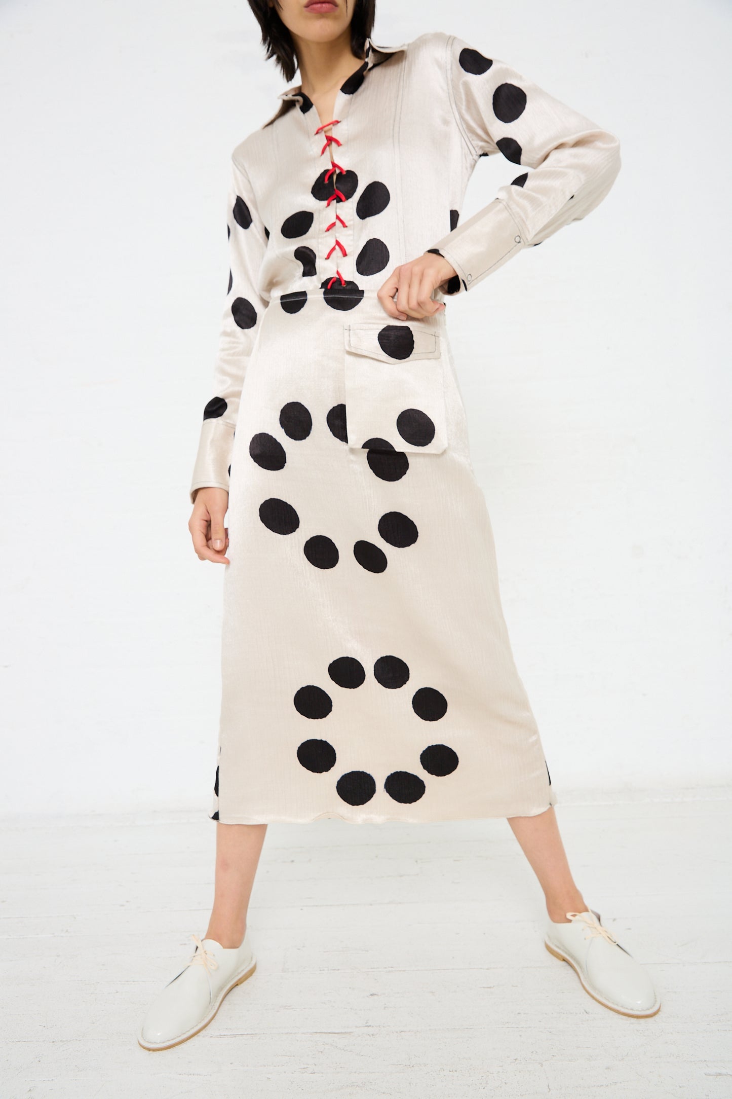 Person wearing a beige Silk Mashroo Midi Skirt by TIGRA TIGRA with large black polka dots and red lace-up detail, paired with white shoes. The skirt, featuring contrast topstitching, has long sleeves and a pocket.