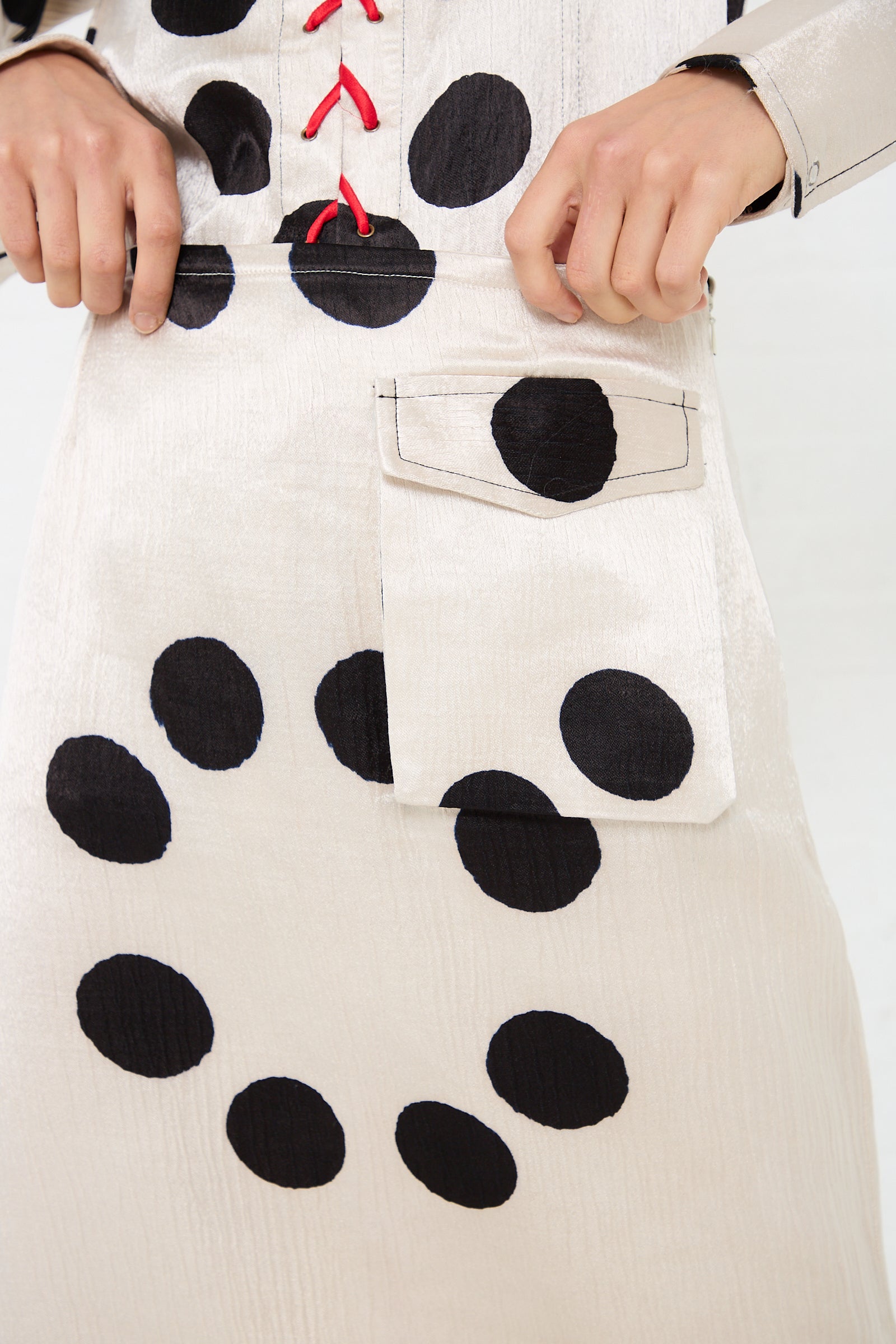 A person adjusts a pocket on a beige garment with large black polka dots. The Silk Mashroo Midi Skirt from TIGRA TIGRA features red lacing on the upper section, adding an elegant touch to its unique design.