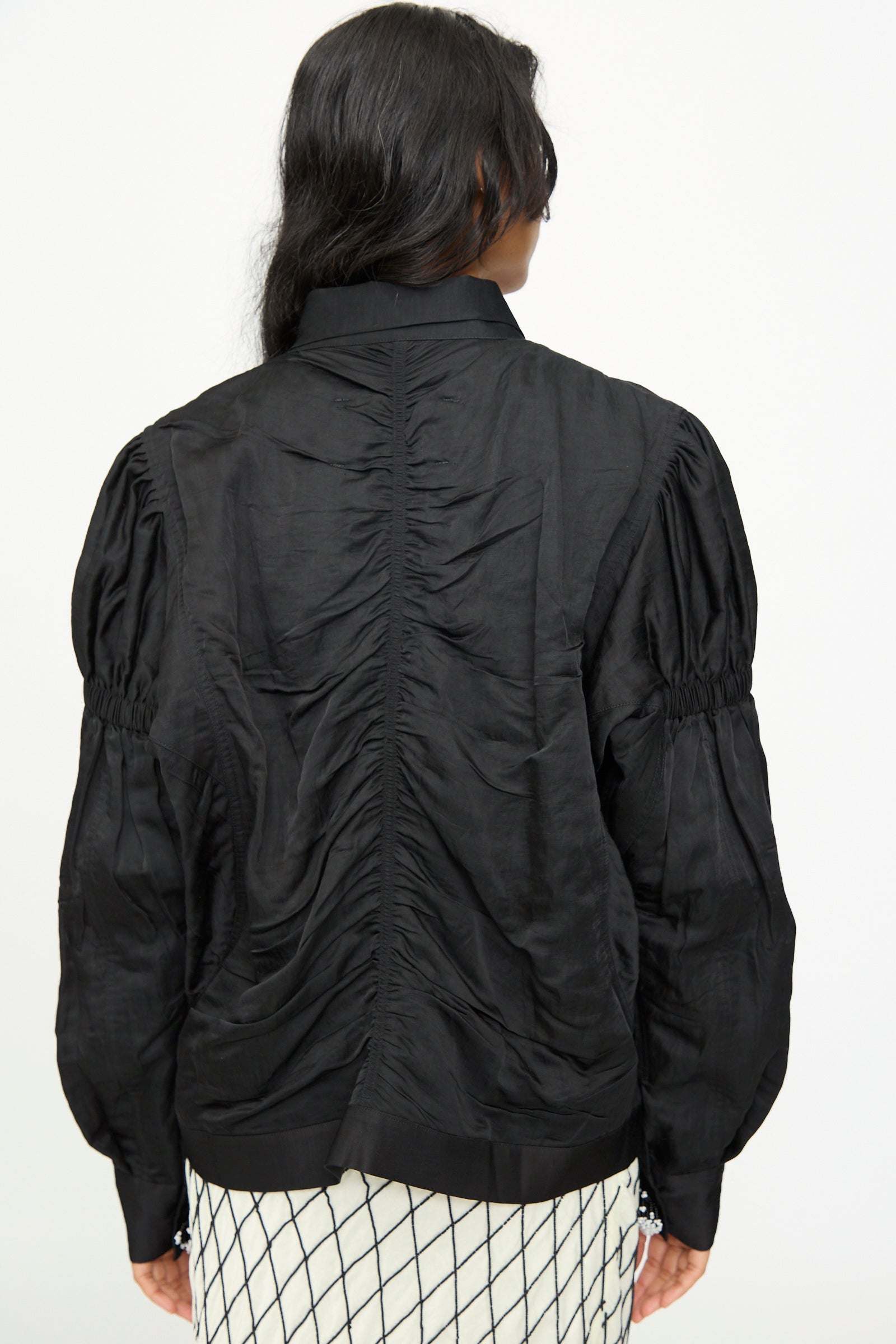 A person with long dark hair is seen from behind wearing a black ruched long-sleeve blouse, a white skirt with a black grid pattern, and the TIGRA TIGRA Victory Bomber with Beaded Buttons.