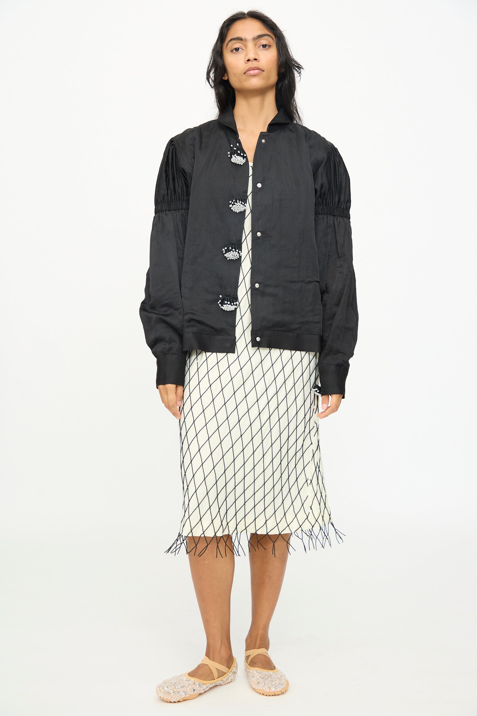 A person stands facing forward, wearing TIGRA TIGRA’s Victory Bomber with Beaded Buttons—a black hand-loomed jacket—over a white dress with a black grid pattern and fringe at the hem, paired with beige woven shoes.