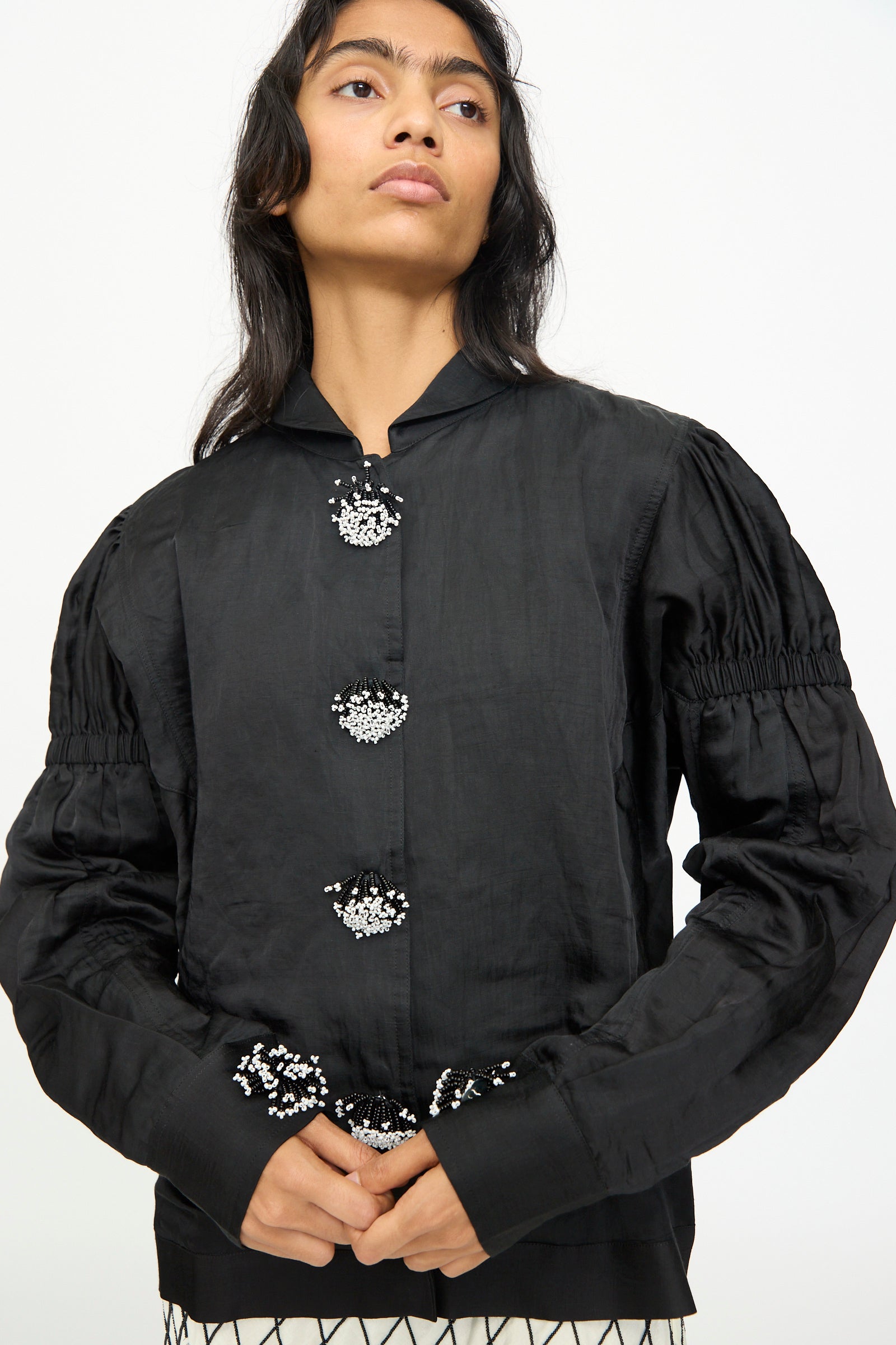 A woman stands against a plain white background wearing the TIGRA TIGRA Victory Bomber—a black long-sleeve blouse featuring decorative beaded buttons, gathered sleeves, and a high collar.