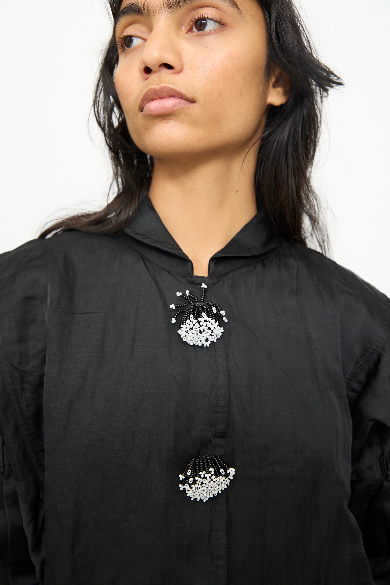 Wearing the TIGRA TIGRA Victory Bomber with Beaded Buttons, a person looks slightly away from the camera. The black, hand-loomed jacket features two beaded floral clasps.