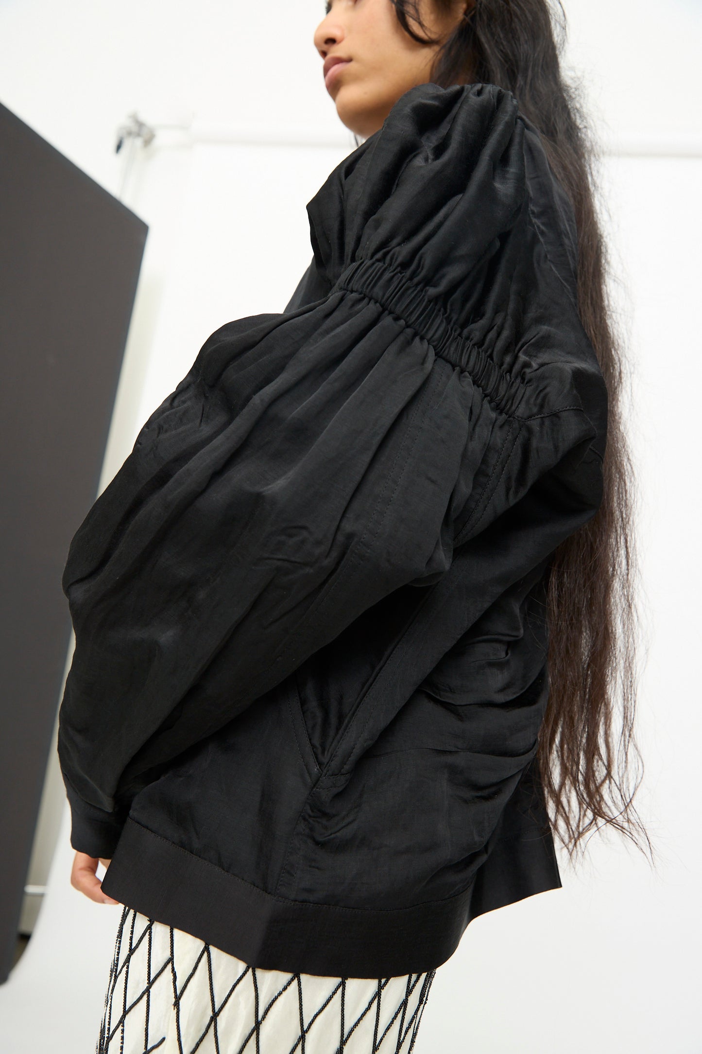 A person stands in a minimalist, white-walled space, wearing a black ruched long-sleeve blouse and a patterned white skirt, with the TIGRA TIGRA Victory Bomber with Beaded Buttons.