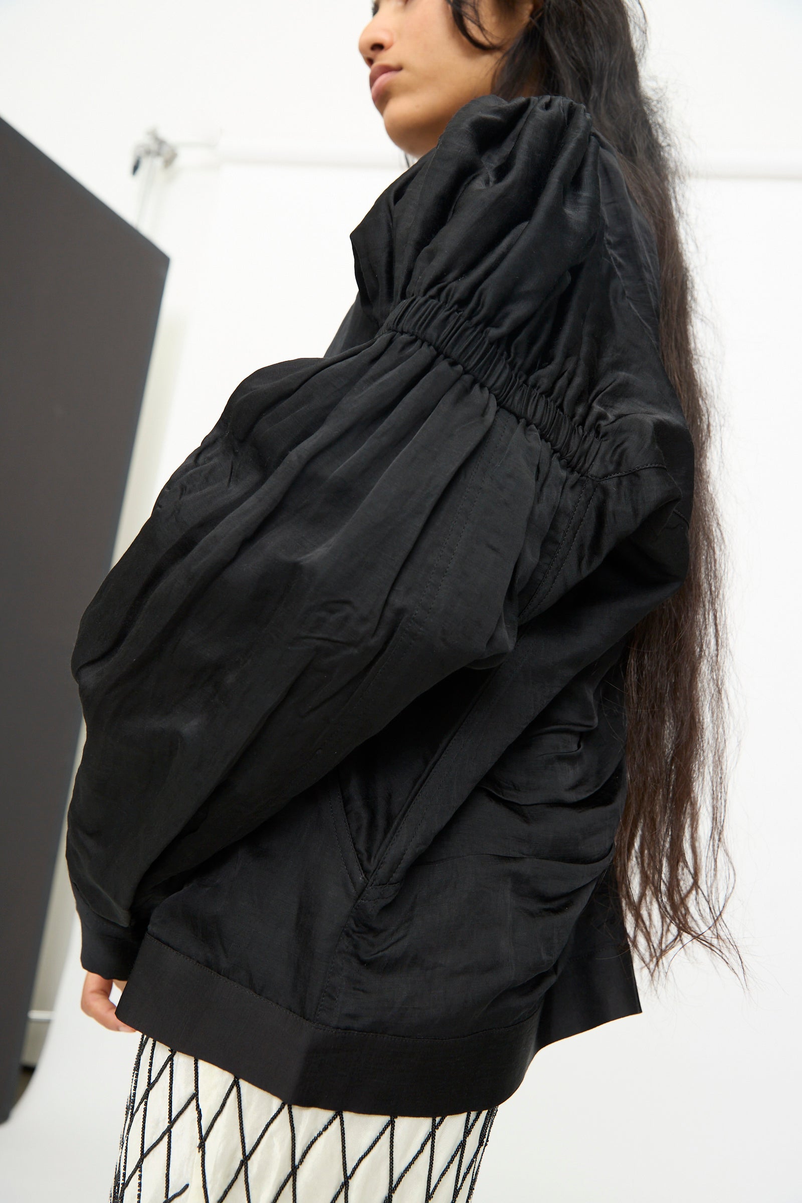 A person stands in a minimalist, white-walled space, wearing a black ruched long-sleeve blouse and a patterned white skirt, with the TIGRA TIGRA Victory Bomber with Beaded Buttons.