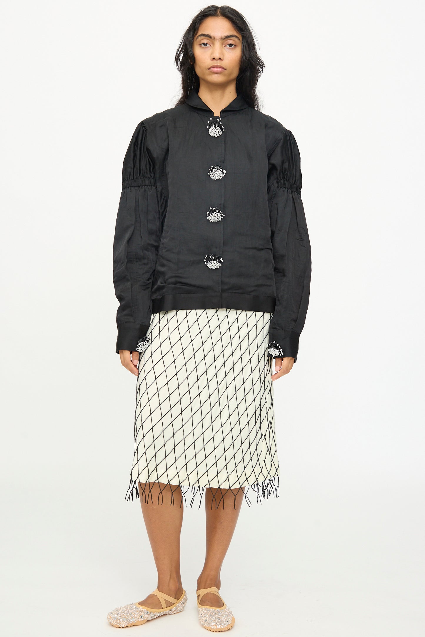 A person wearing the TIGRA TIGRA Victory Bomber with Beaded Buttons paired with a white skirt featuring a black net-like pattern, standing against a plain white background.