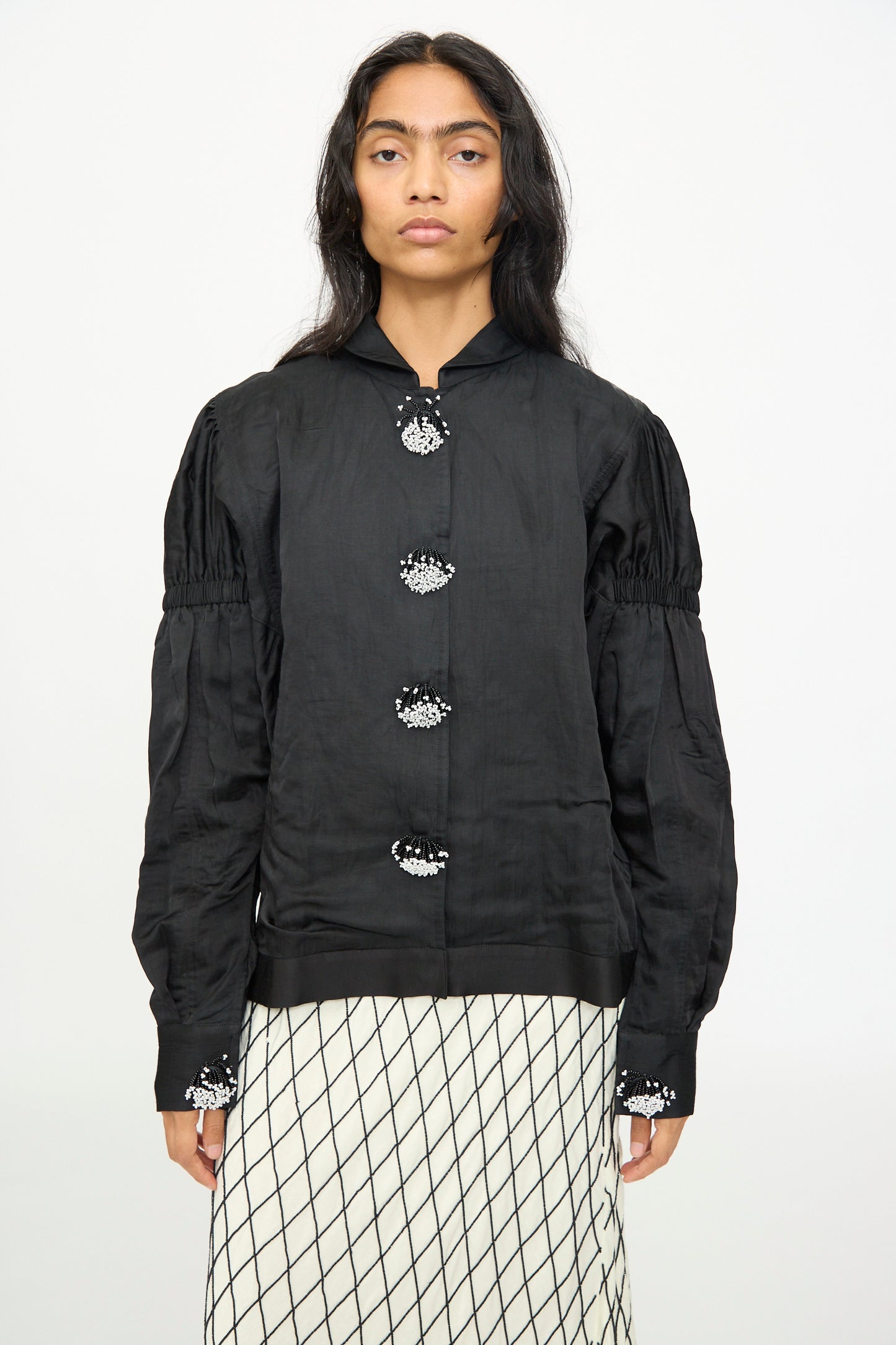 A model wears the TIGRA TIGRA Victory Bomber with Beaded Buttons—a black hand-loomed jacket with gathered sleeves—paired with a white skirt featuring a black grid pattern, standing against a plain background.
