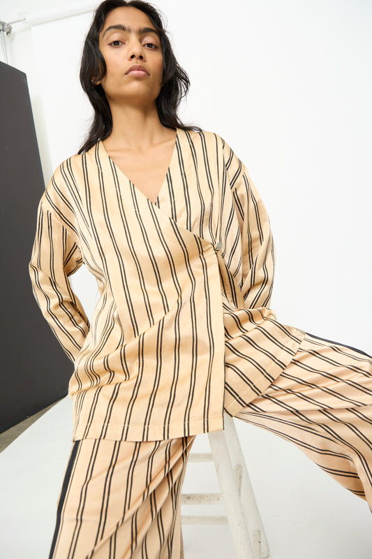 A person wearing the TIGRA TIGRA Block Printed Striped Wrap Top in Champagne and Black sits on a white stool against a plain background, looking at the camera. 